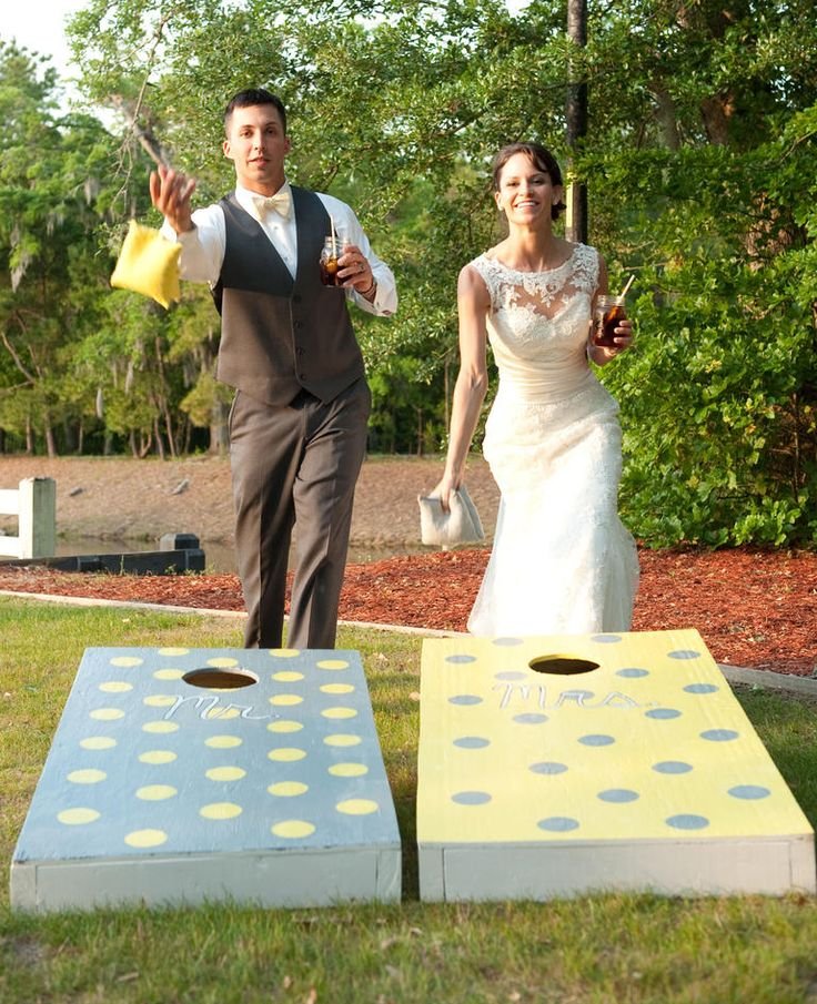 A newlywed couple playing the game of cornhole outdoors. The groom is tossing a yellow bean bag while holding a drink, and the bride is holding a drink and a white clutch. They are standing behind two cornhole boards labeled 'Mr.' and 'Mrs.' with polka dot patterns, on a grassy area with trees in the background.