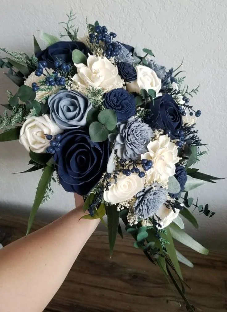 Hand holding a bouquet of artificial flowers with blue, white, and gray roses, green leaves, and small blue berries against a plain wall background.