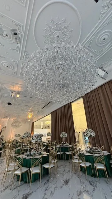 Elegant event hall decorated with a large crystal chandelier, round tables with green tablecloths, gold chairs, and floral centerpieces, with brown curtains and marble flooring.