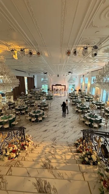 Elegant banquet hall decorated for a wedding reception with chandeliers, round tables set with white tablecloths and chairs, a stage with floral garland backdrop, and a grand staircase decorated with flowers.