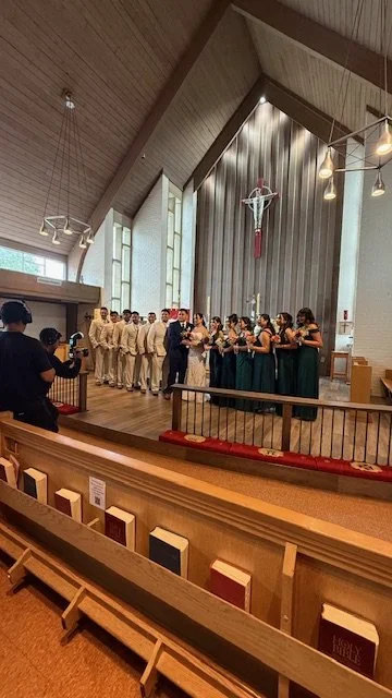 A group of people dressed in formal attire gathered inside a church, with some holding flowers, during a ceremony near the altar with a crucifix hanging on the wall behind them.