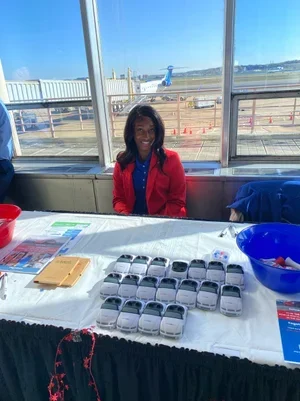 A woman smiling at a table with small white car models displayed in front of her at an airport terminal. There are various bowls and pamphlets on the table, and aircraft are visible outside the window.