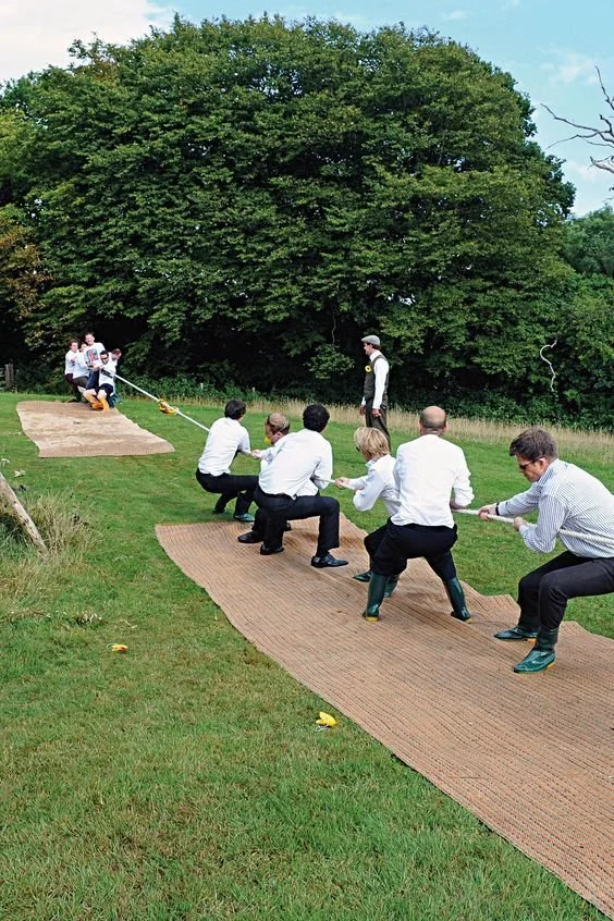 People participating in a tug-of-war game outdoors on grass, with a large tree and open sky in the background.