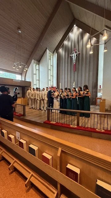 A wedding ceremony taking place inside a church with the bride, groom, bridesmaids, and groomsmen standing at the altar, with the church's interior and large cross visible in the background.