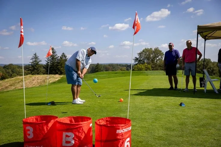 People playing golf on a bright, sunny day with a golf course and trees in the background, two people standing under a tent watching, and golf bags and flags on the green.