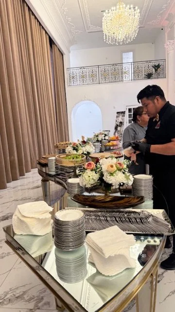People serving food at a buffet table decorated with flowers in an elegant room with a chandelier and marble floors.