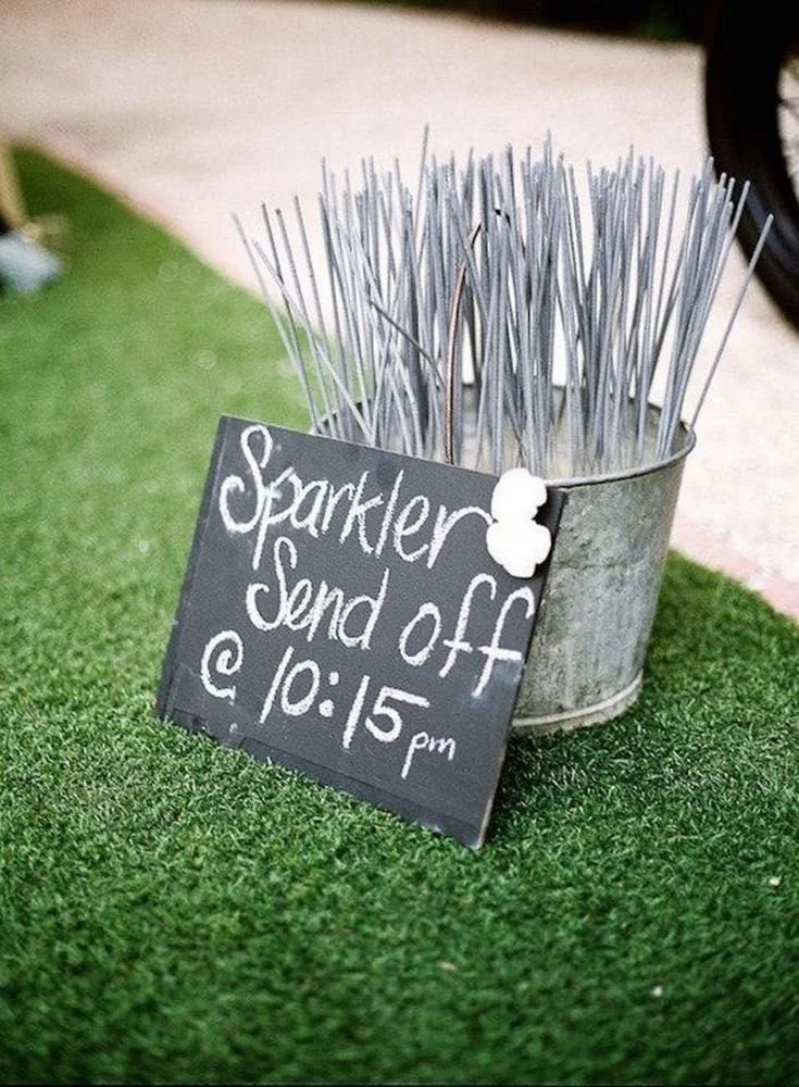 A metal bucket filled with toothpicks, placed on artificial grass. A small blackboard sign in front reads 'Sparkler Send off @ 10:15 pm'.