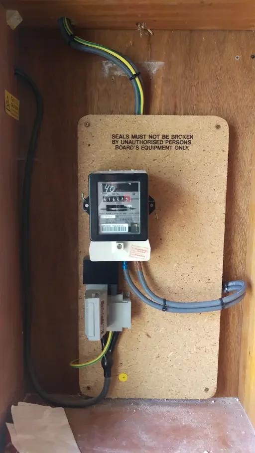 An electrical meter mounted on a wooden board inside a wooden enclosure, with various electrical wires connected to it. There is a warning printed on the wooden board stating, "Seals must not be broken by unauthorized persons. Board's equipment only.