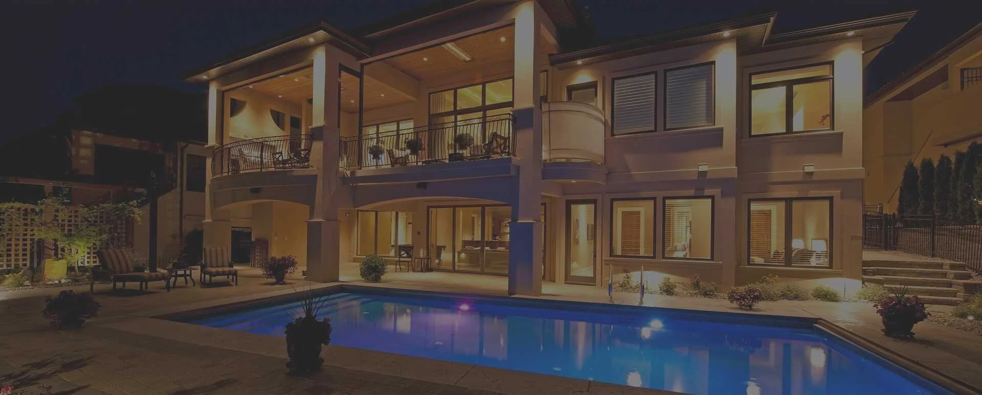 Night view of a modern multi-story house with illuminated interior and exterior, a backyard swimming pool, patio furniture, potted plants, and a staircase leading to an upper level.