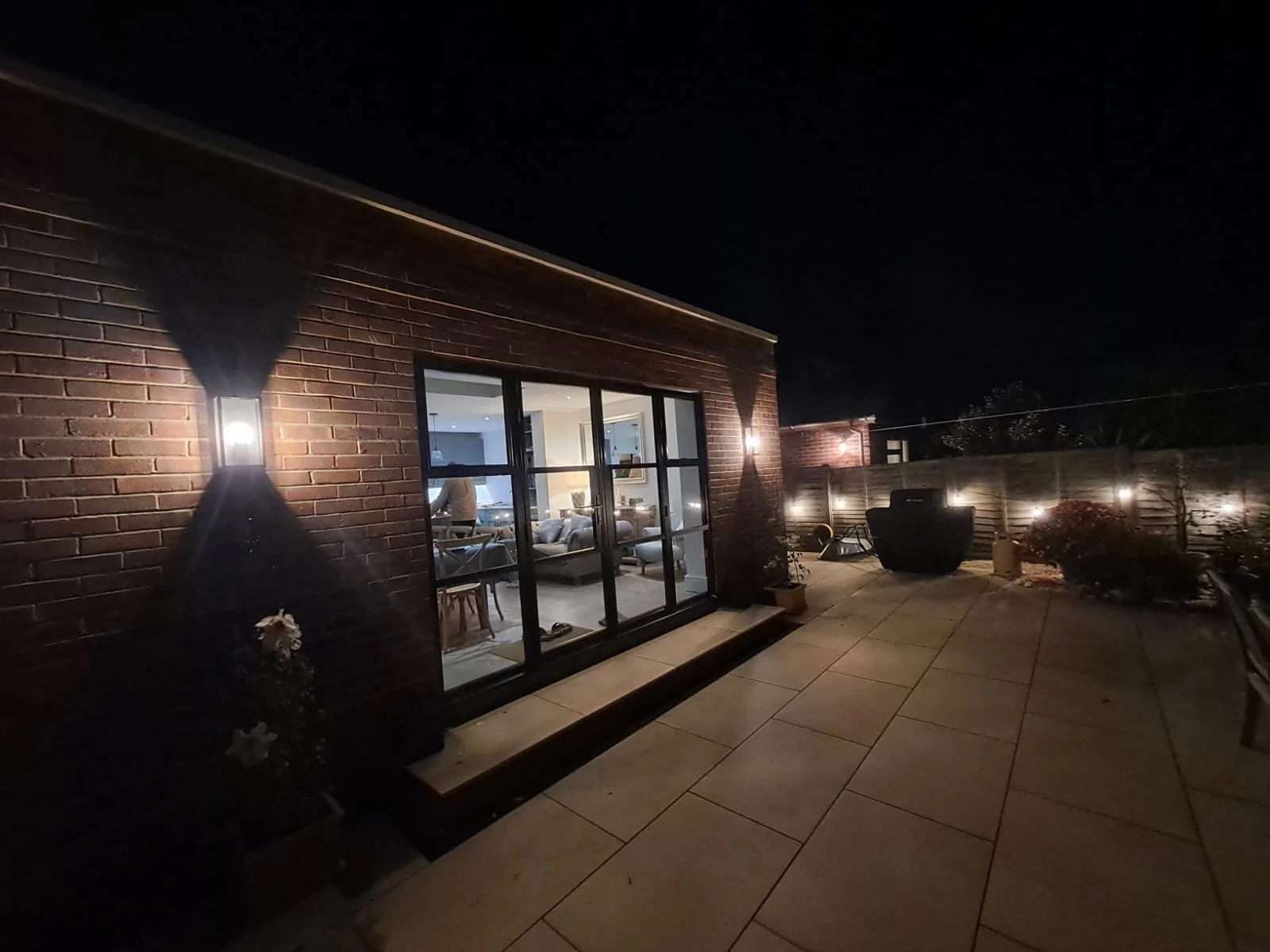 Night time view of a brick house with lit outdoor wall sconces, large glass doors, and a spacious patio with chairs and plants.