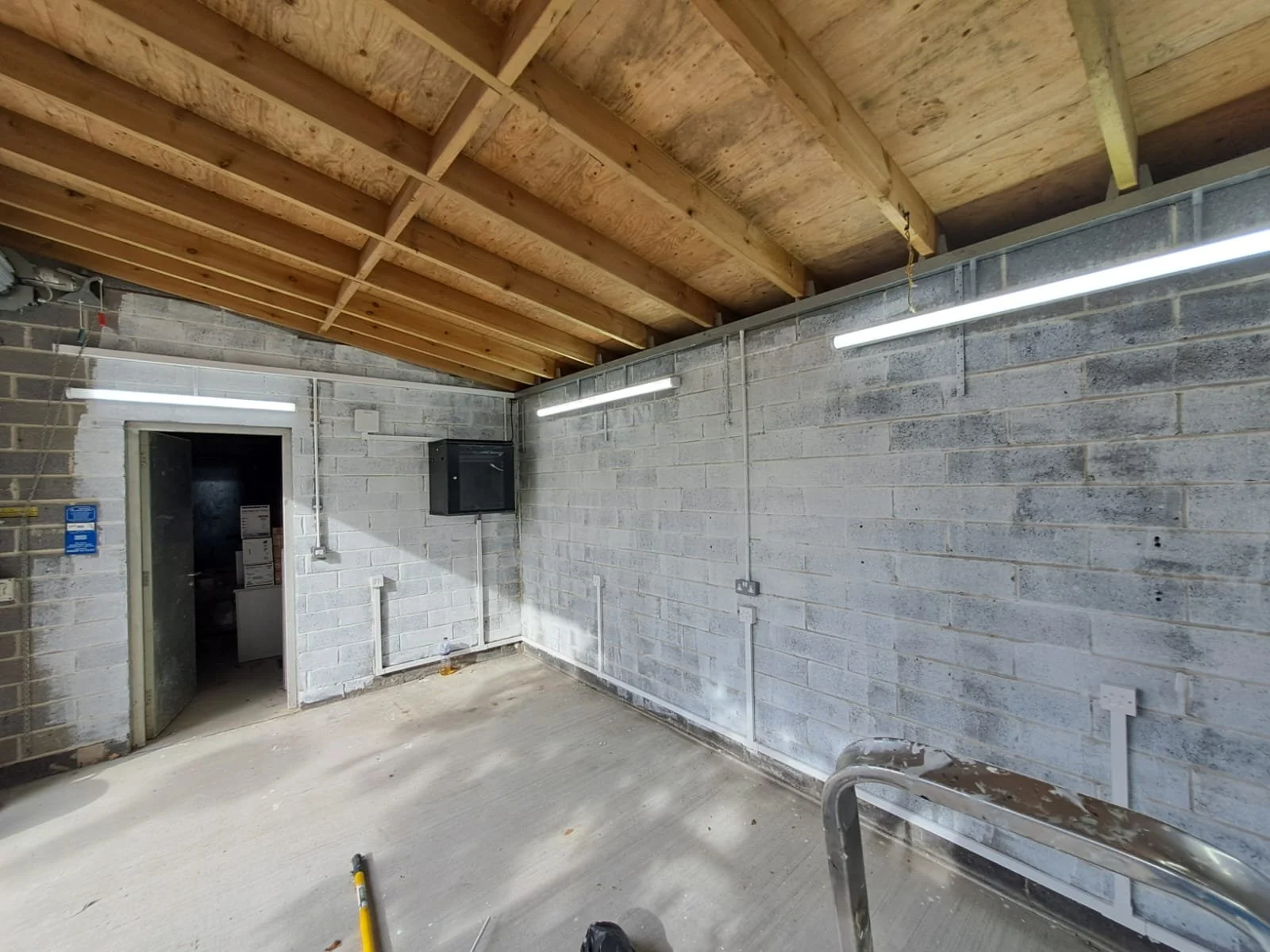 Empty industrial or commercial room with unfinished concrete floors, cinder block walls, and a wooden ceiling with exposed beams. Fluorescent lights are mounted on the walls, and there are electrical outlets and a black box on the wall.