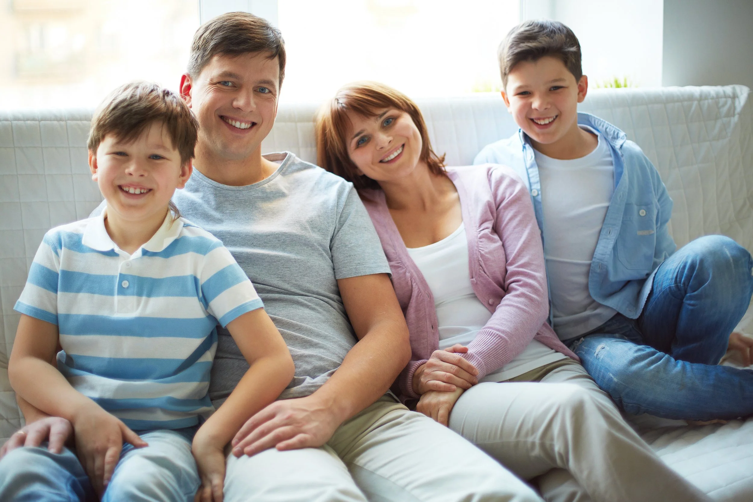 A family of five sitting on a white couch and smiling at the camera. The family includes two boys, a woman, a man, and a girl, in a bright living room.