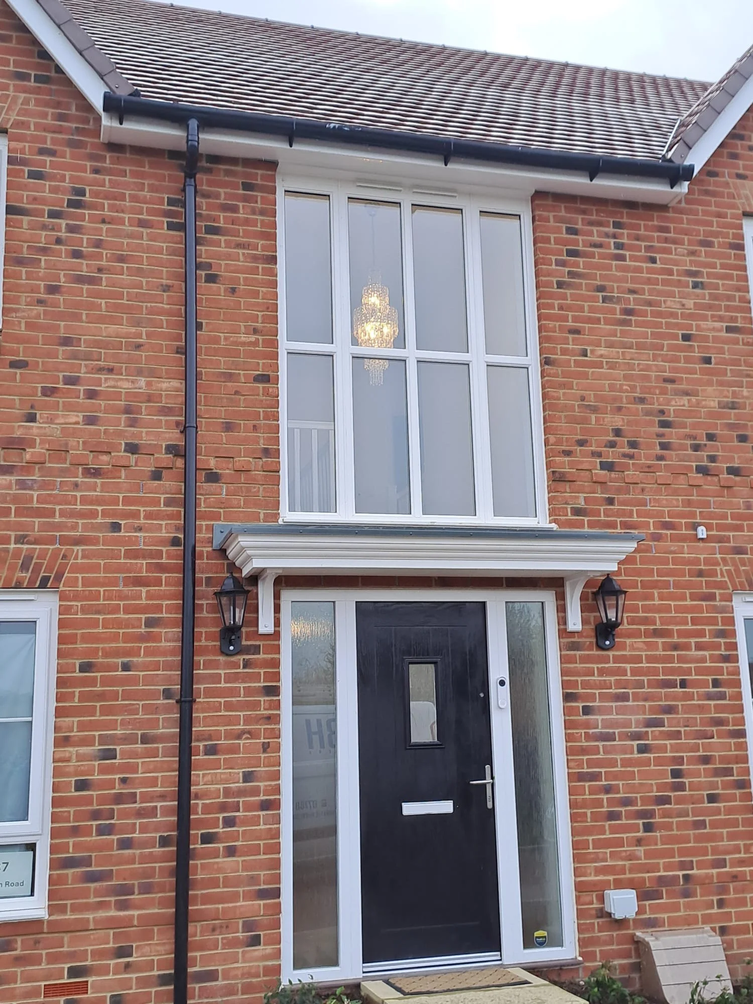 The front of a brick house with a black door, two black wall-mounted lanterns, and a large glass window with a chandelier visible inside.