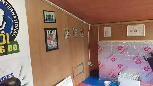 Indoor room with wooden walls, framed pictures, electrical wiring, and a mattress wrapped in plastic, with a fan and heater.
