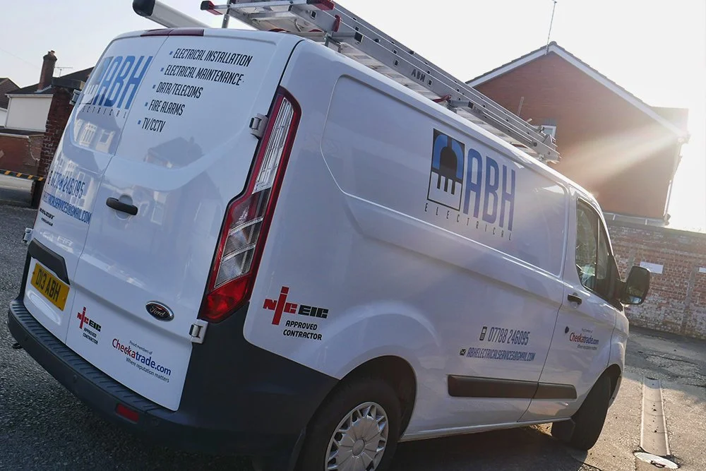 A white commercial van with company branding and contact information parked on the street. The van has a ladder on top and text indicating electrical services.