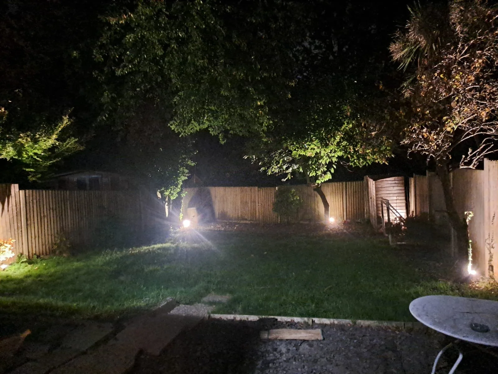 Night time backyard with trees, a lawn, a concrete patio, a wooden privacy fence, sidewalk, garden lights, and a white patio table.