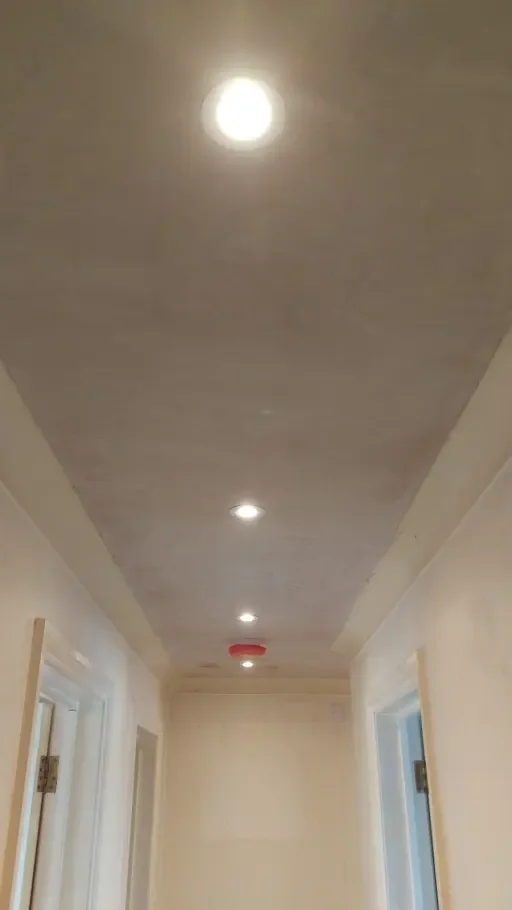 View of a hallway ceiling with three recessed lights and a smoke detector, with part of a door frame visible at each side.
