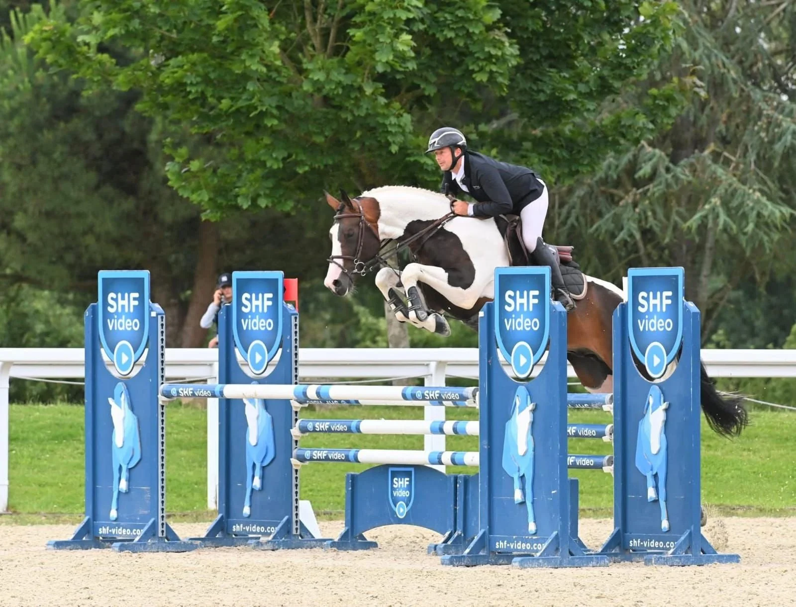 Un cavalier en compétition de saut d'obstacles, sautant une barrière bleue avec des graphismes SHF vidéo, dans un environnement extérieur avec des arbres en arrière-plan.