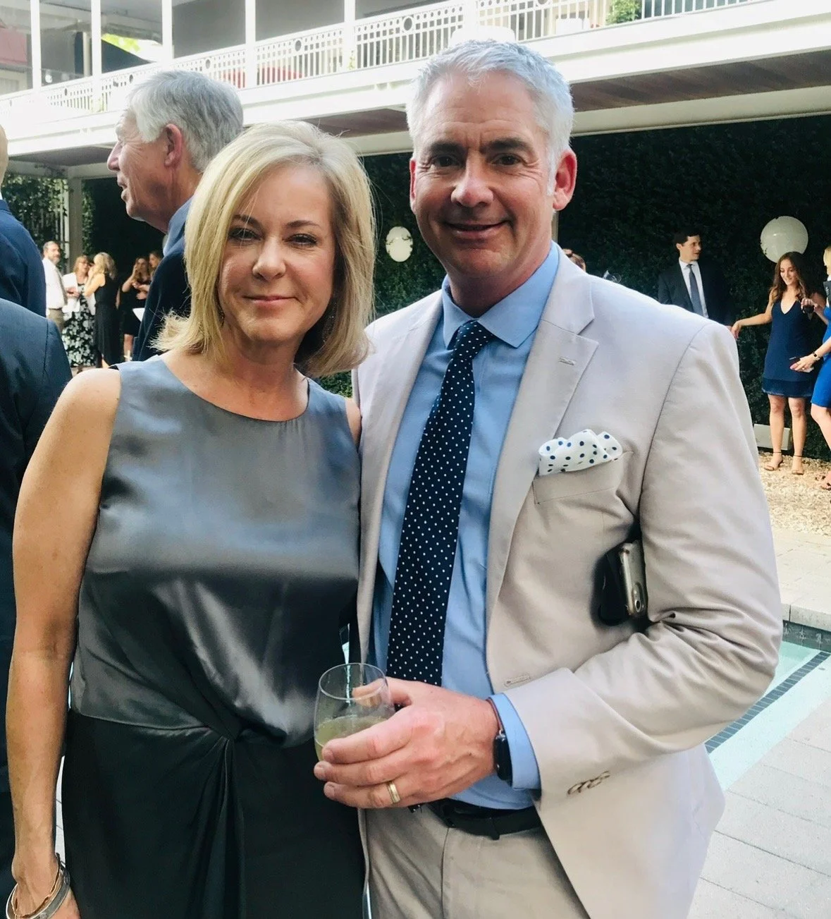 A woman and a man dressed in formal attire at an outdoor event. The woman has shoulder-length blonde hair and is wearing a sleeveless gray satin dress. The man has short gray hair, is wearing a light-colored suit with a blue shirt, polka-dot tie, and pocket square. He is holding a glass of white wine.