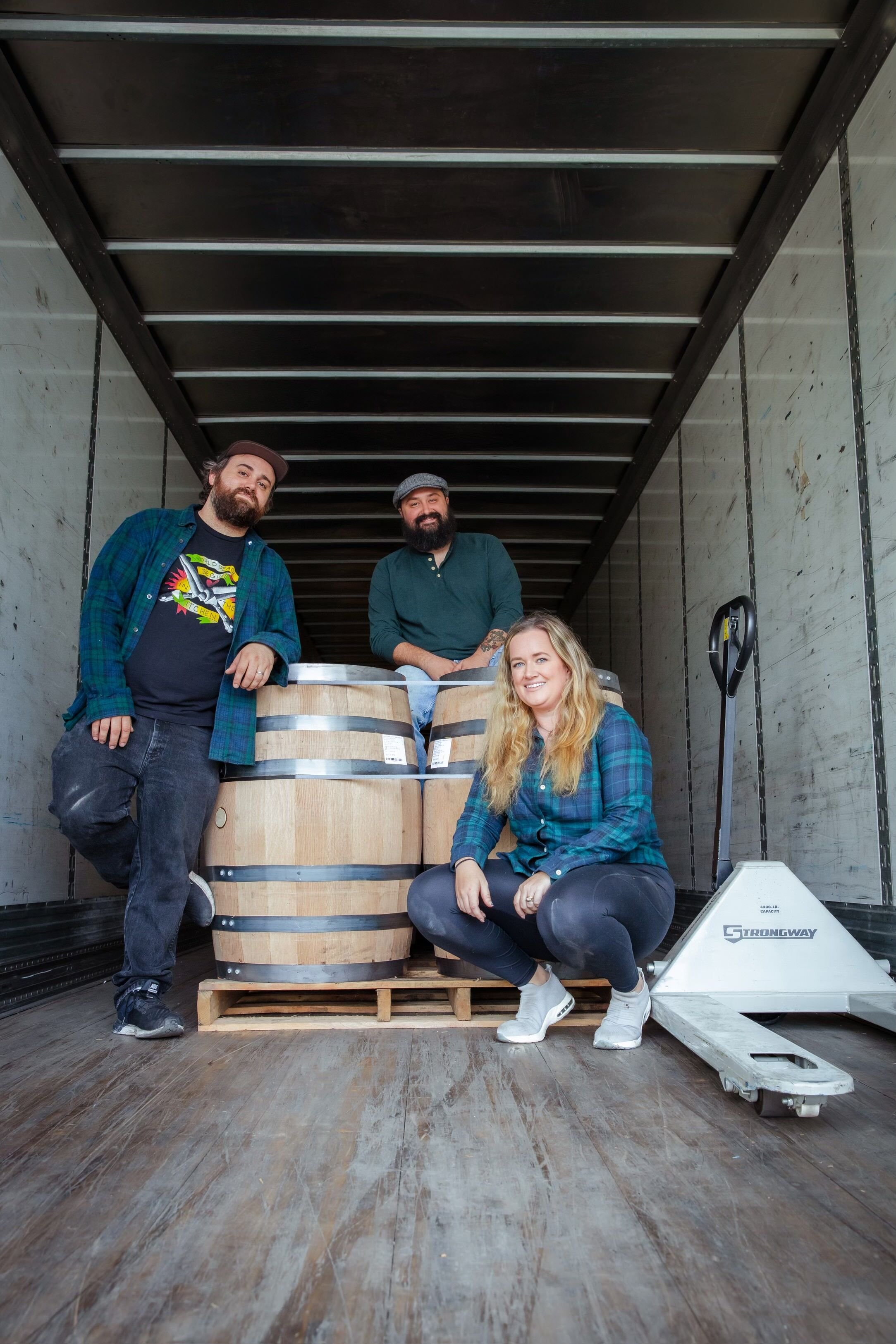 Two men stand inside a brewery, holding glasses of beer and toasting, surrounded by large stainless steel fermentation tanks.
