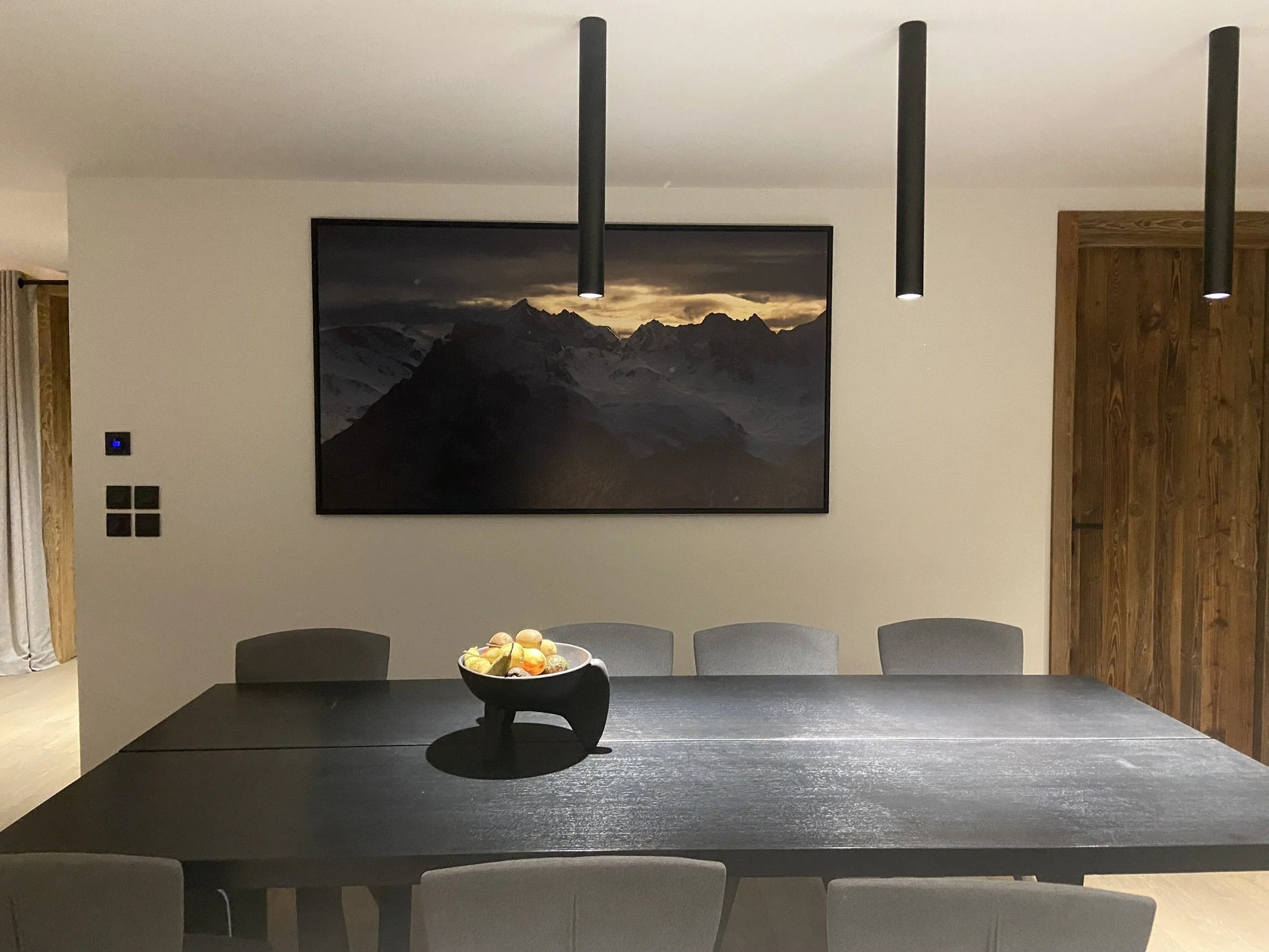 Salle à manger avec une grande table, un tableau représentant des montagnes et un centre de table avec des fruits.