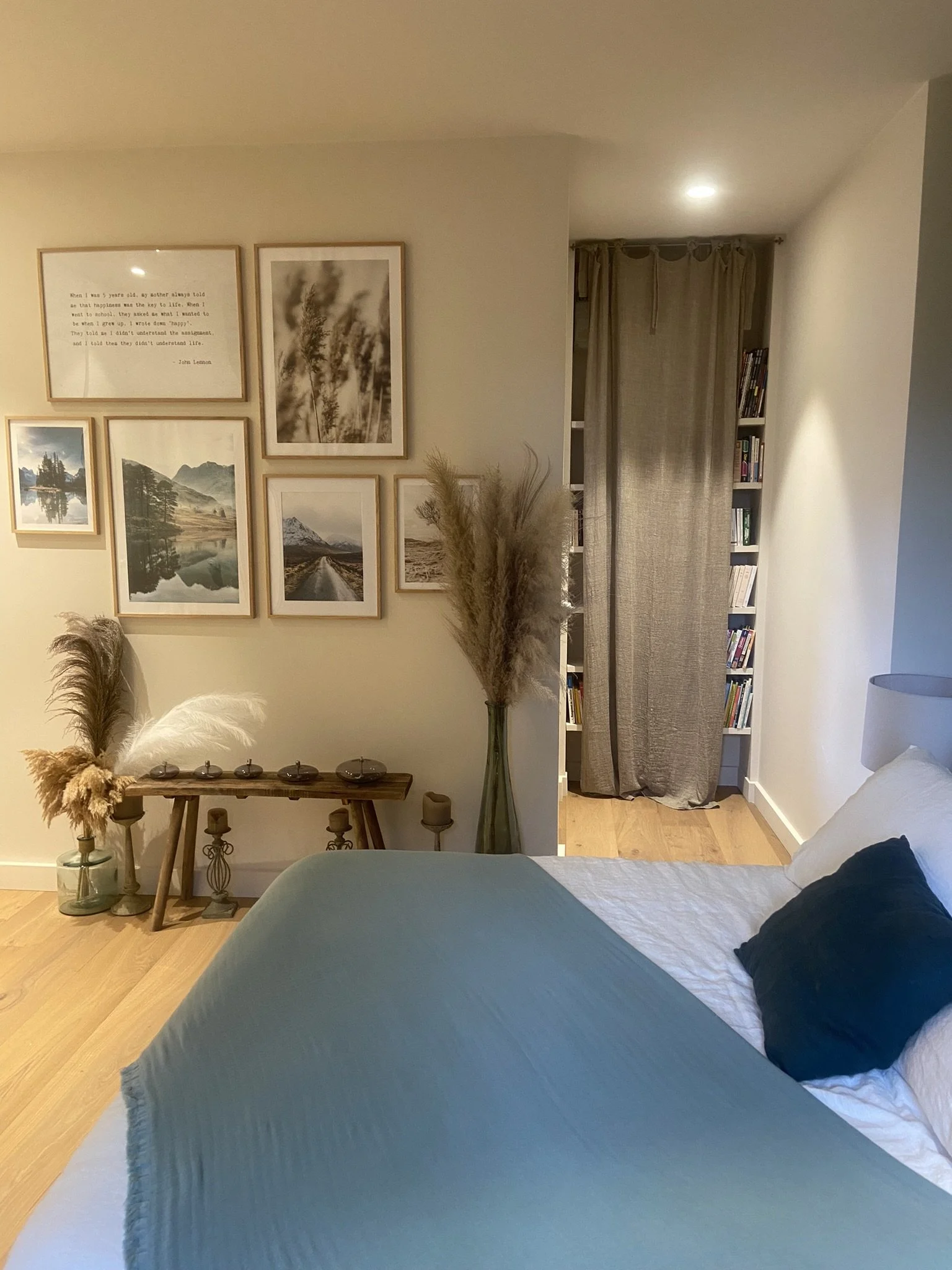 Chambre décorée dans un style farmhouse avec un lit, des coussins, un vase avec des plantes séchées, une étagère avec des livres, et un mur avec des cadres photo et une citation.