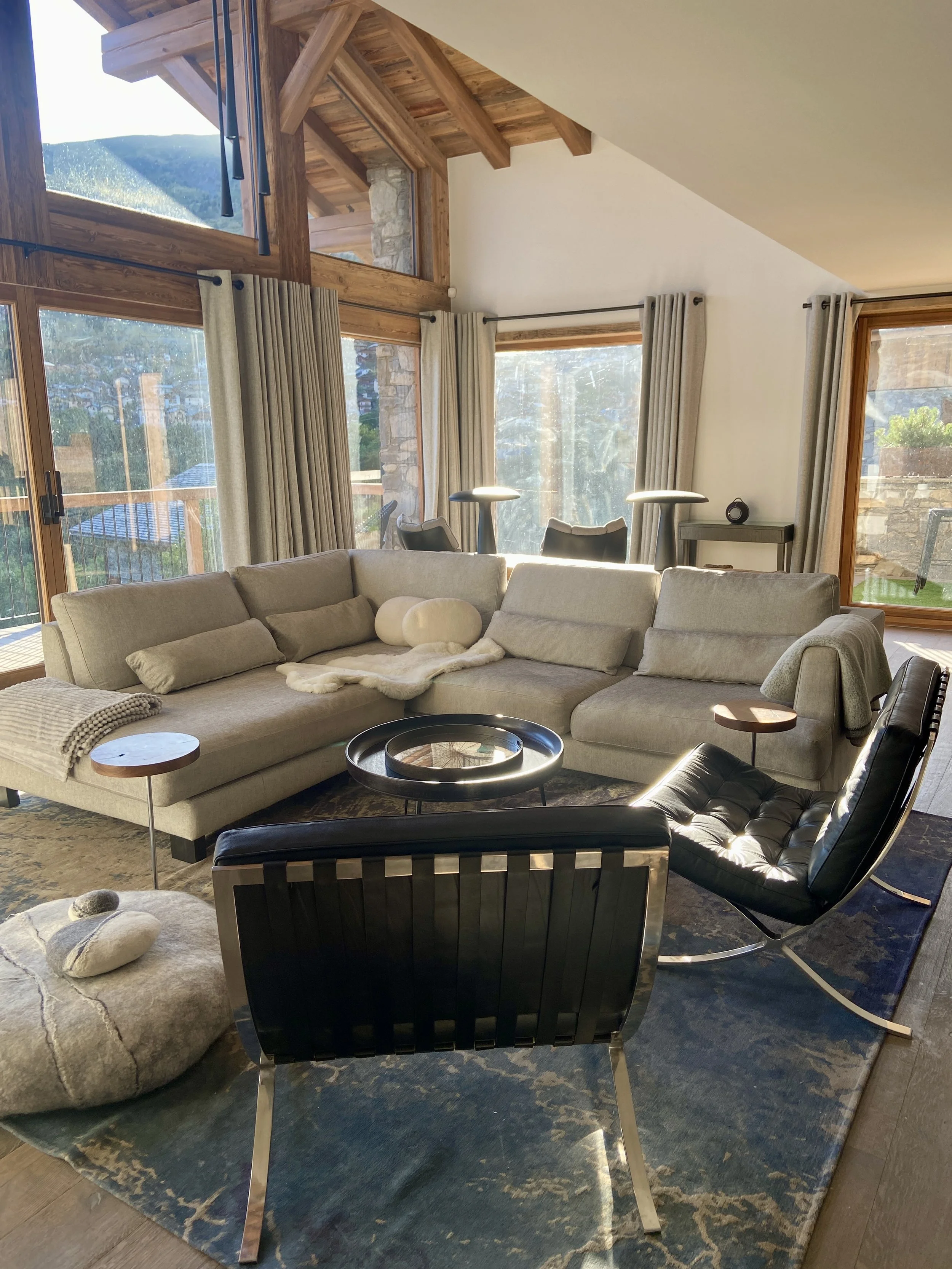 Salon de salon avec canapé beige, chaises en cuir noir, table basse ronde, grande fenêtre avec vue sur la nature, rideaux beiges, plafond en bois, tapis bleu et beige, décor moderne