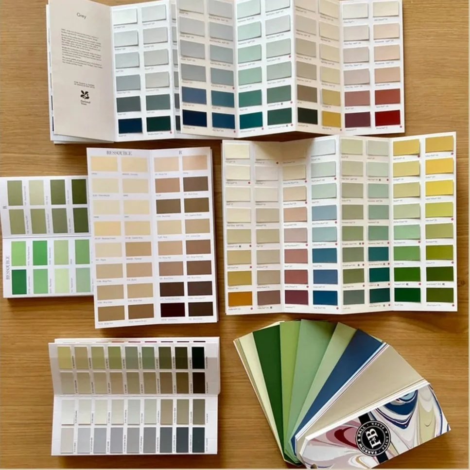 Échantillonneur de couleurs avec plusieurs nuanciers ouverts et un échantillonneur stylé avec différentes nuances de vert, beige, et gris sur une surface en bois.