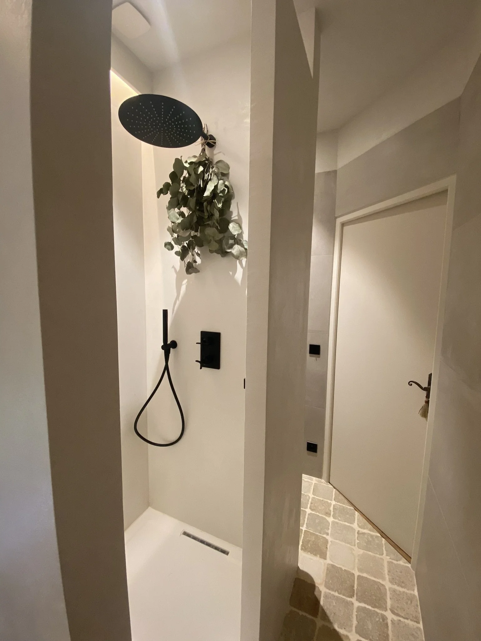 Une douche moderne avec un plafond de pluie noir, une pomme de douche noire, un rideau de douche noir, un mur décoré de plantes sèches vertes, et un sol en carreaux de pierre. Il y a une porte blanche à droite et quelques prises électriques noires sur le mur.
