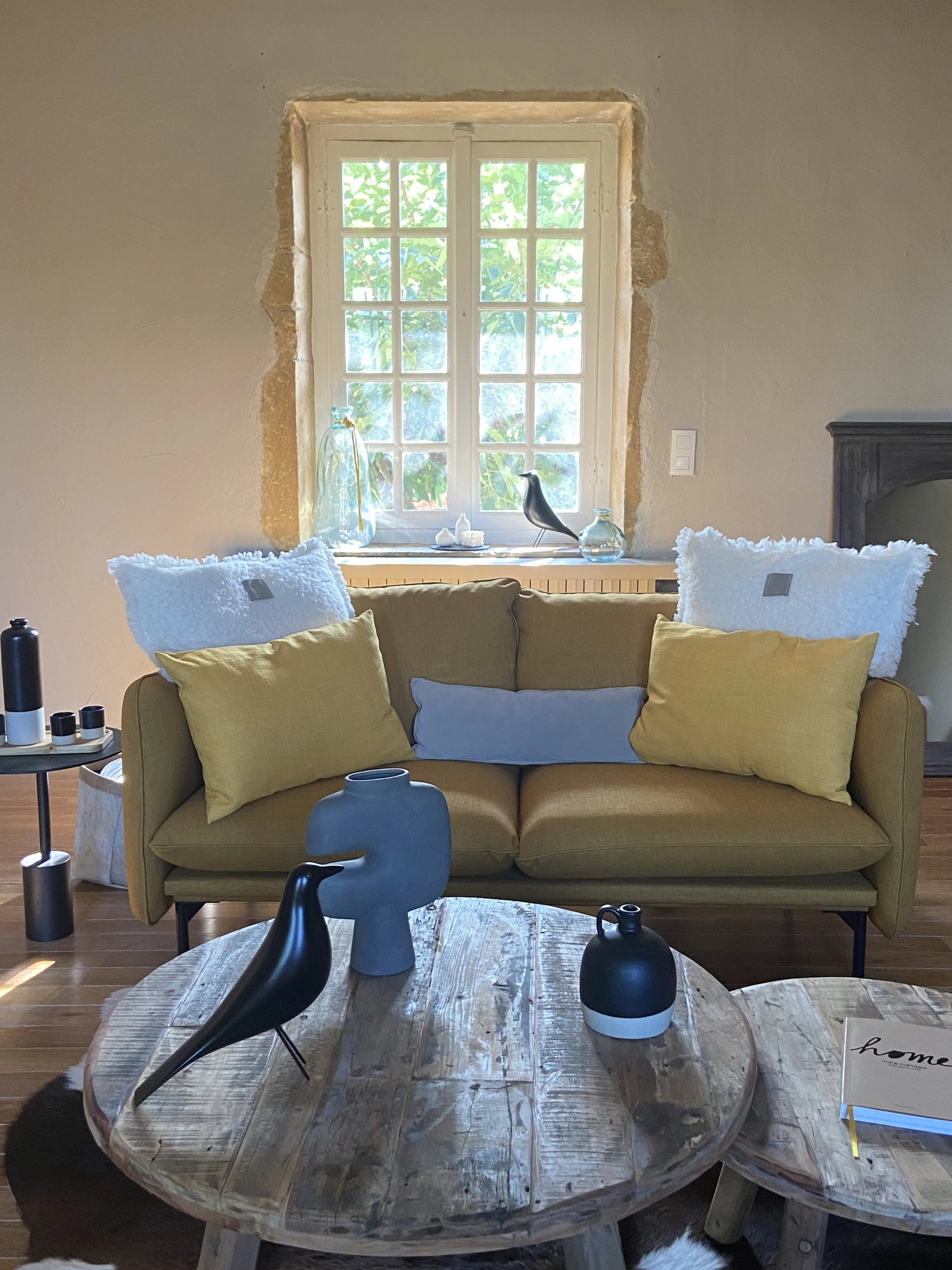 Salon avec canapé jaune, table en bois, décorations en céramique et en verre, fenêtre avec vue sur la nature.