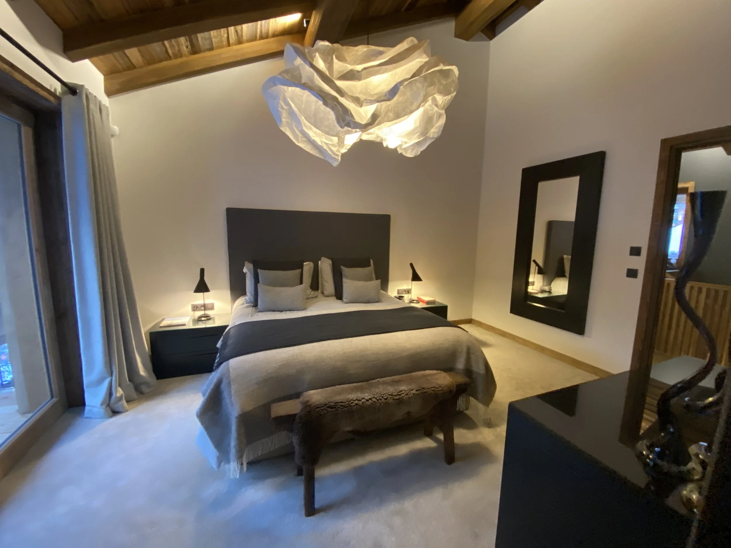 Chambre moderne avec lit double, trois coussins, deux lampes de chevet, miroir noir rectangulaire, décoration en bois au plafond, grande fenêtre avec rideaux, mobilier noir