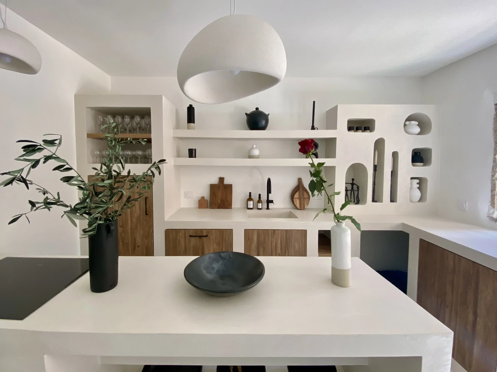 Cuisine minimaliste méditerranéenne moderne avec comptoir blanc, étagères blanches décoratives, vaisselle en verre, vases avec des plantes, et meubles en bois.