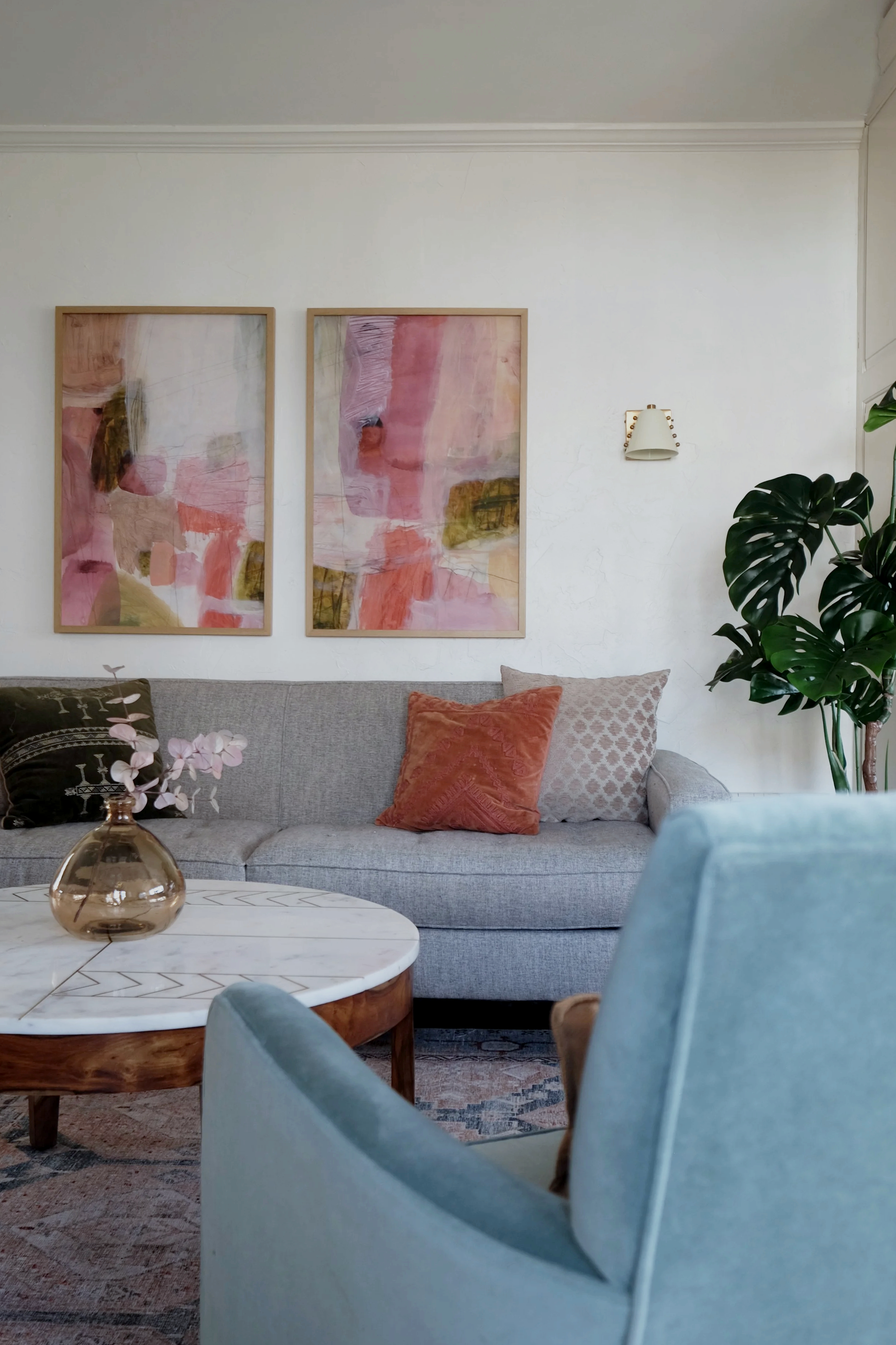 A cozy living room with a gray sofa adorned with decorative pillows, two framed abstract paintings on the wall, a white wall-mounted lamp, a green plant, and a white marble coffee table with a bronze vase and pink flowers.