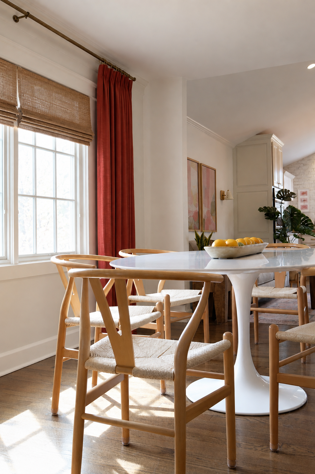 Round white dining table surrounded by wishbone chairs in a sunlit breakfast nook with rust curtains