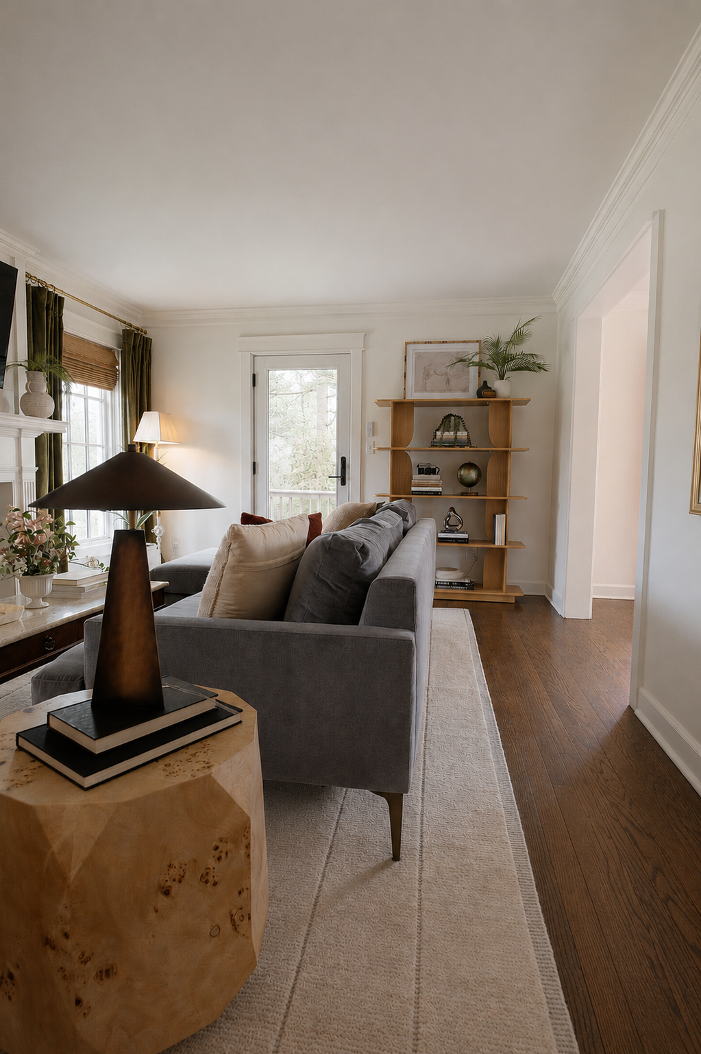 Living room with gray sofa, wooden bookshelf, beige area rug, side table with lamp, and windows with green curtains