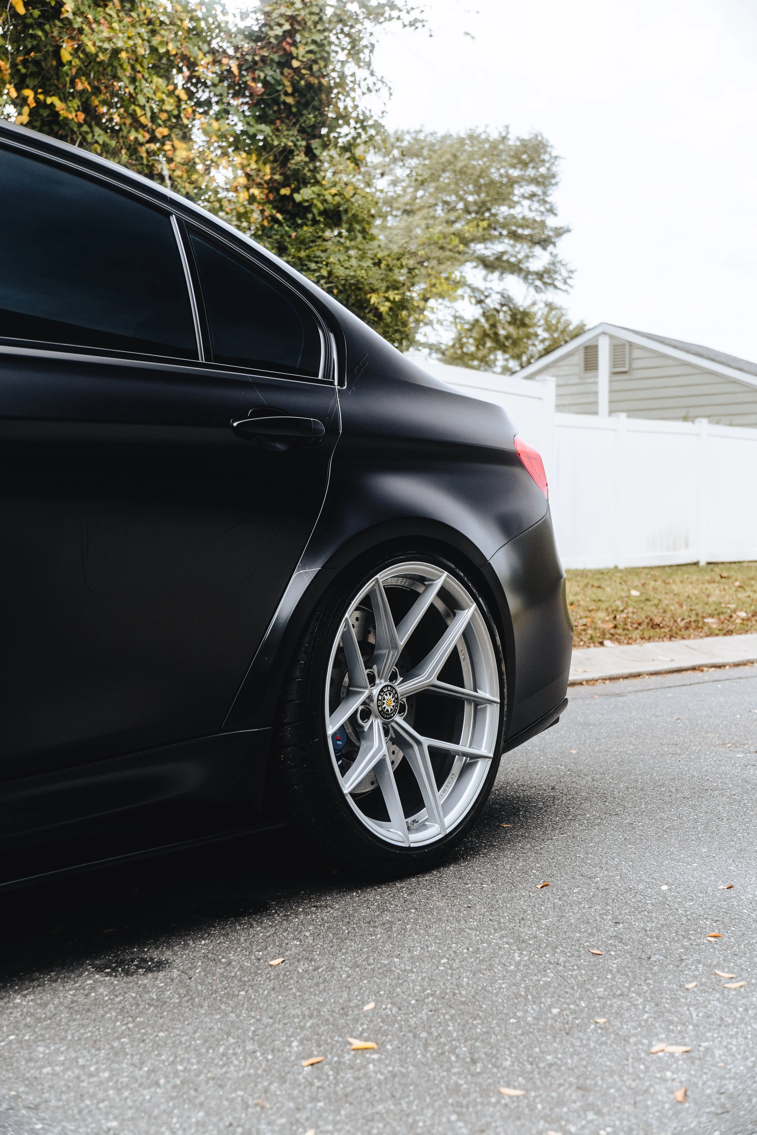 Close-up of the rear side of a black luxury sedan with silver alloy wheels parked on a street with a white fence and trees in the background.