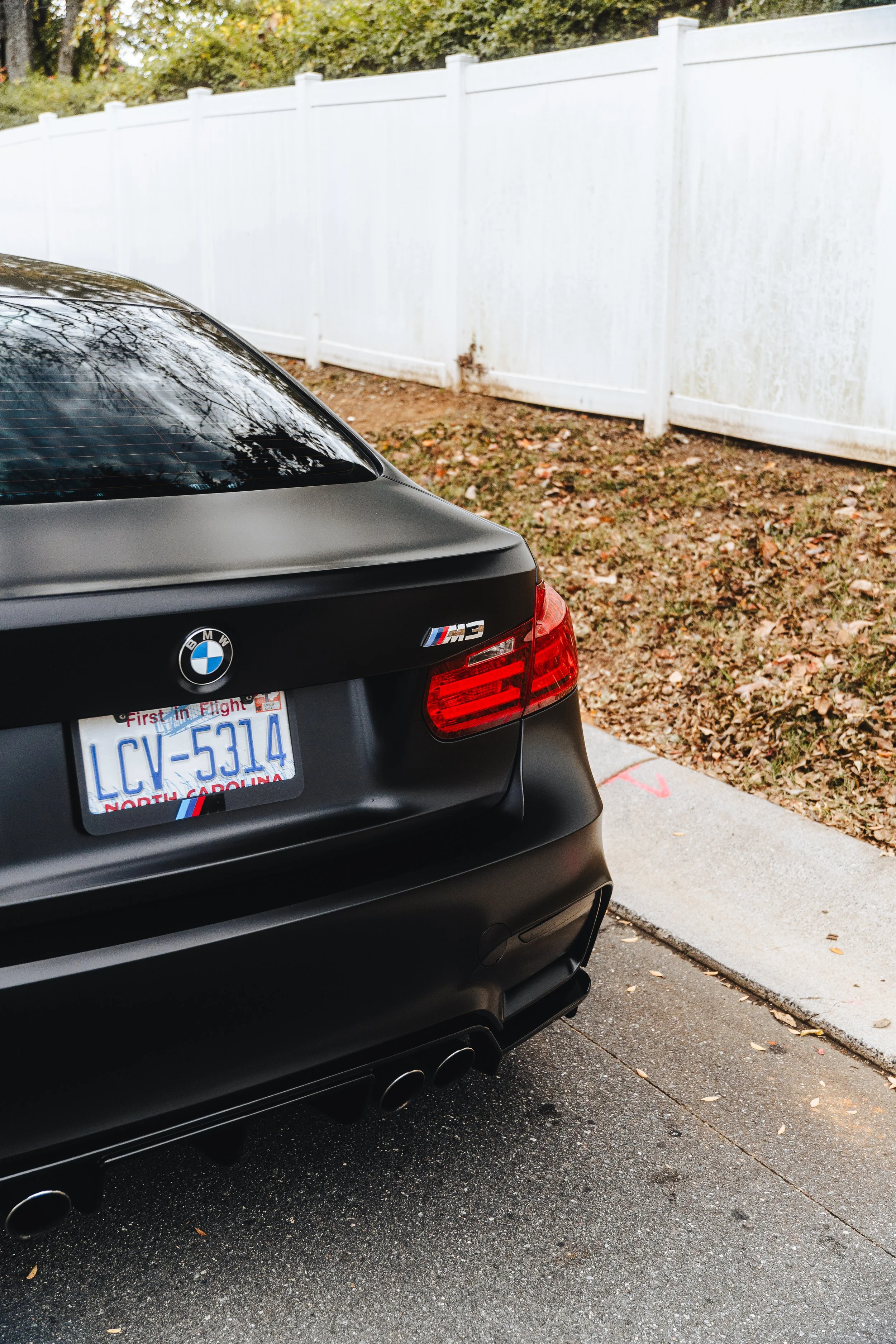 Close-up of the rear of a black BMW M3 parked on a street, showing the rear license plate from North Carolina, taillights, and quad exhaust pipes, with a white fence and some fallen leaves in the background.