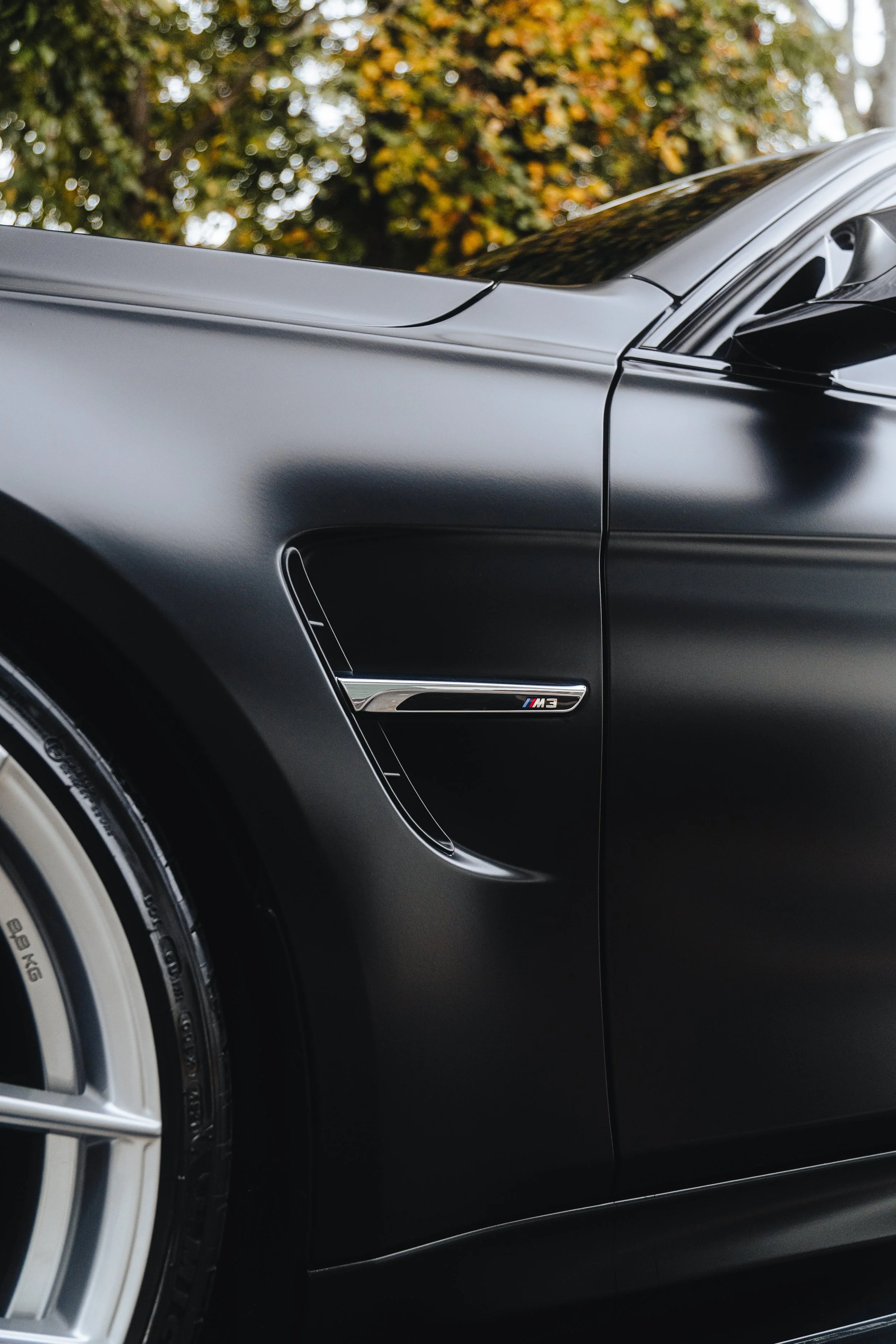Close-up of the front side of a black BMW M3 with matte finish, showing the wheel, vent, and M3 badge.