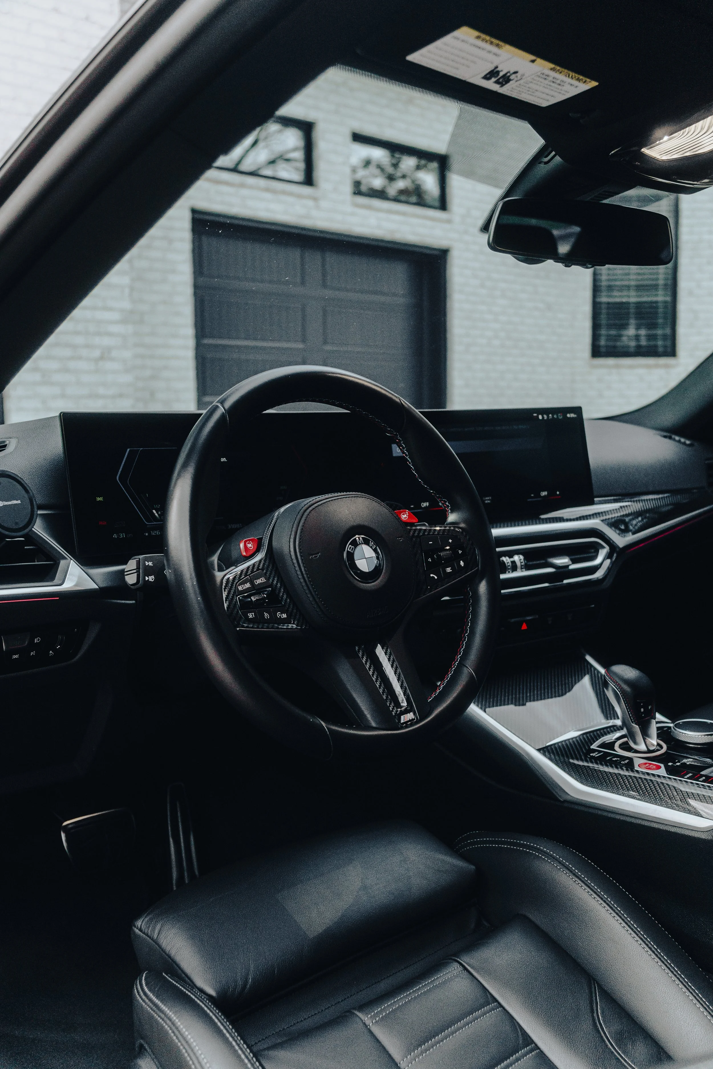 Interior of a BMW car showing the steering wheel, dashboard, infotainment display, gear shift, and black leather seat with white stitching, parked outside a modern white brick house with large windows and a black garage door.
