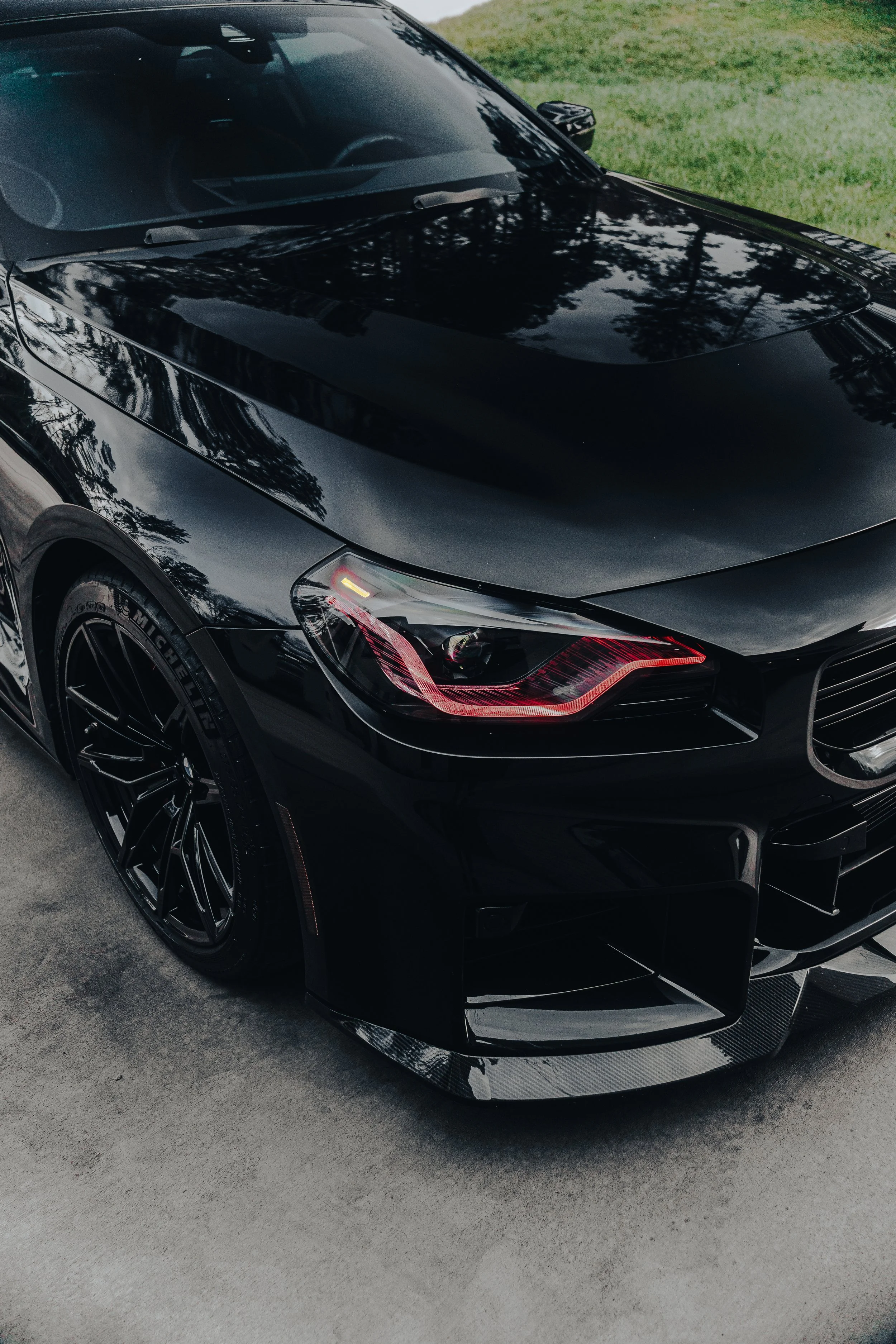 Close-up of a sleek black sports car with tinted windows, modern headlight design, black alloy wheels, and reflective body, parked on a concrete driveway with green grass nearby.