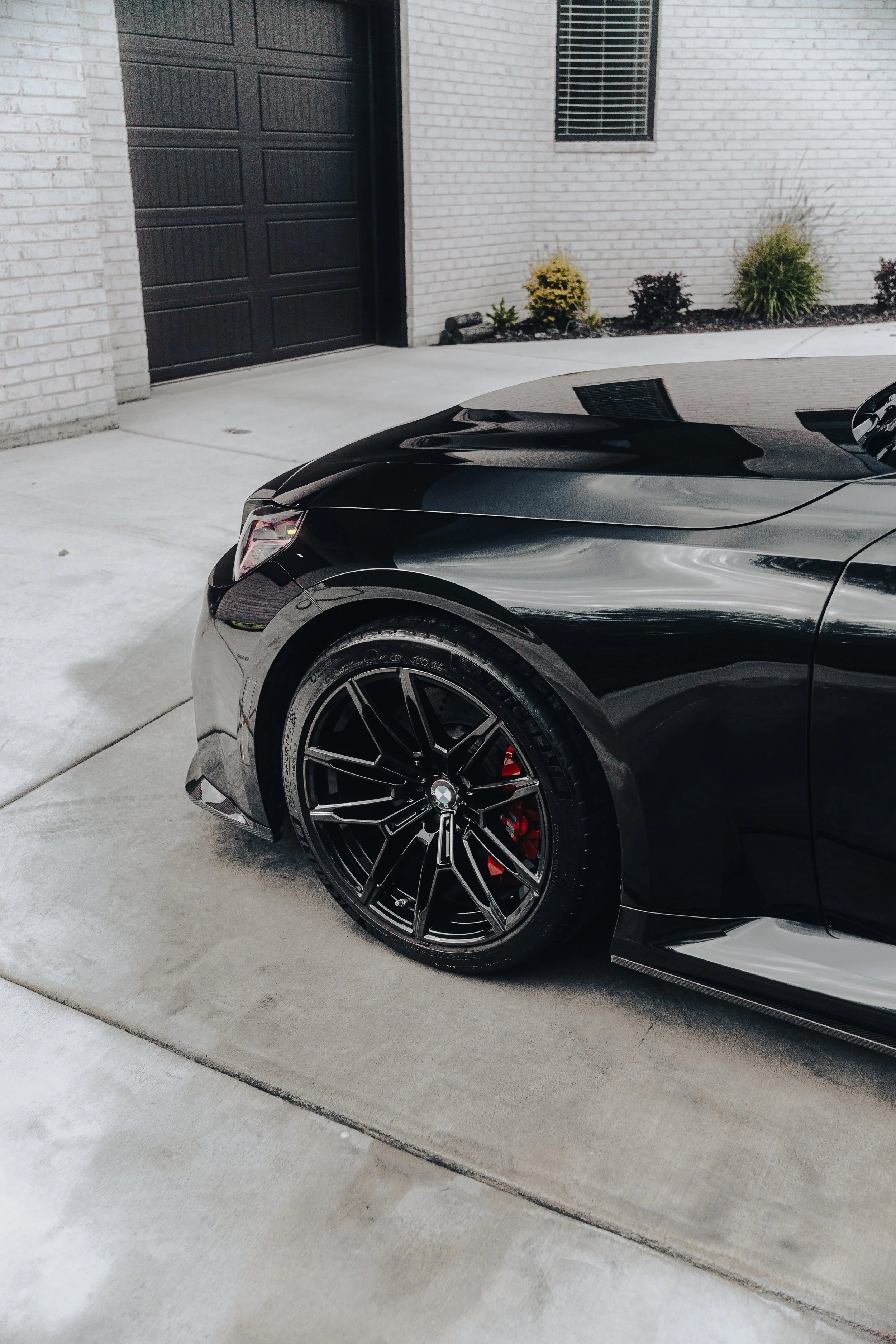 Close-up of the front part of a black BMW sports car parked in a driveway in front of a white brick house with a large black garage door, a window, and small landscaped plants.