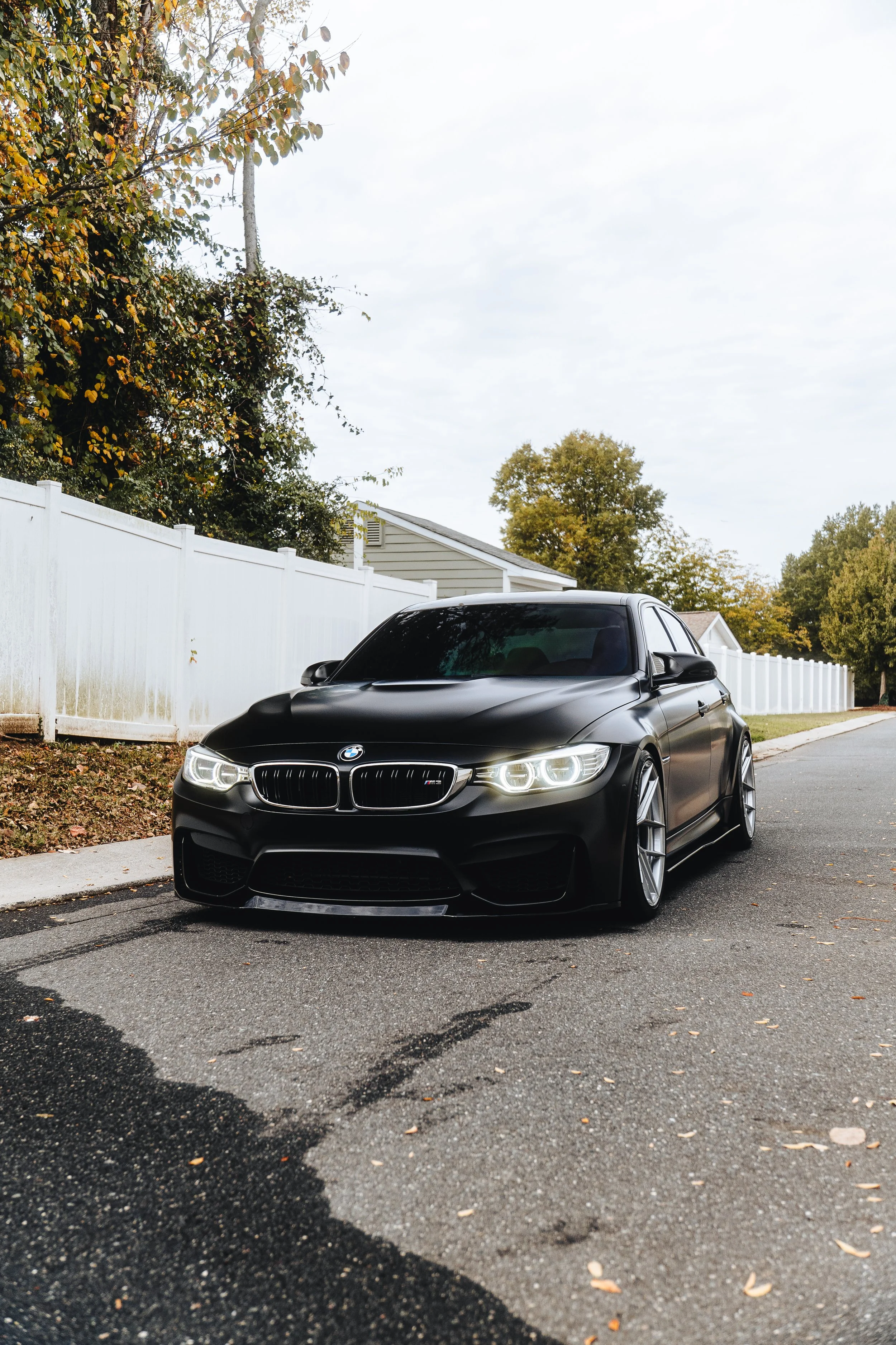 A black BMW M series car parked on the street near a white fence, with trees in the background and an overcast sky.