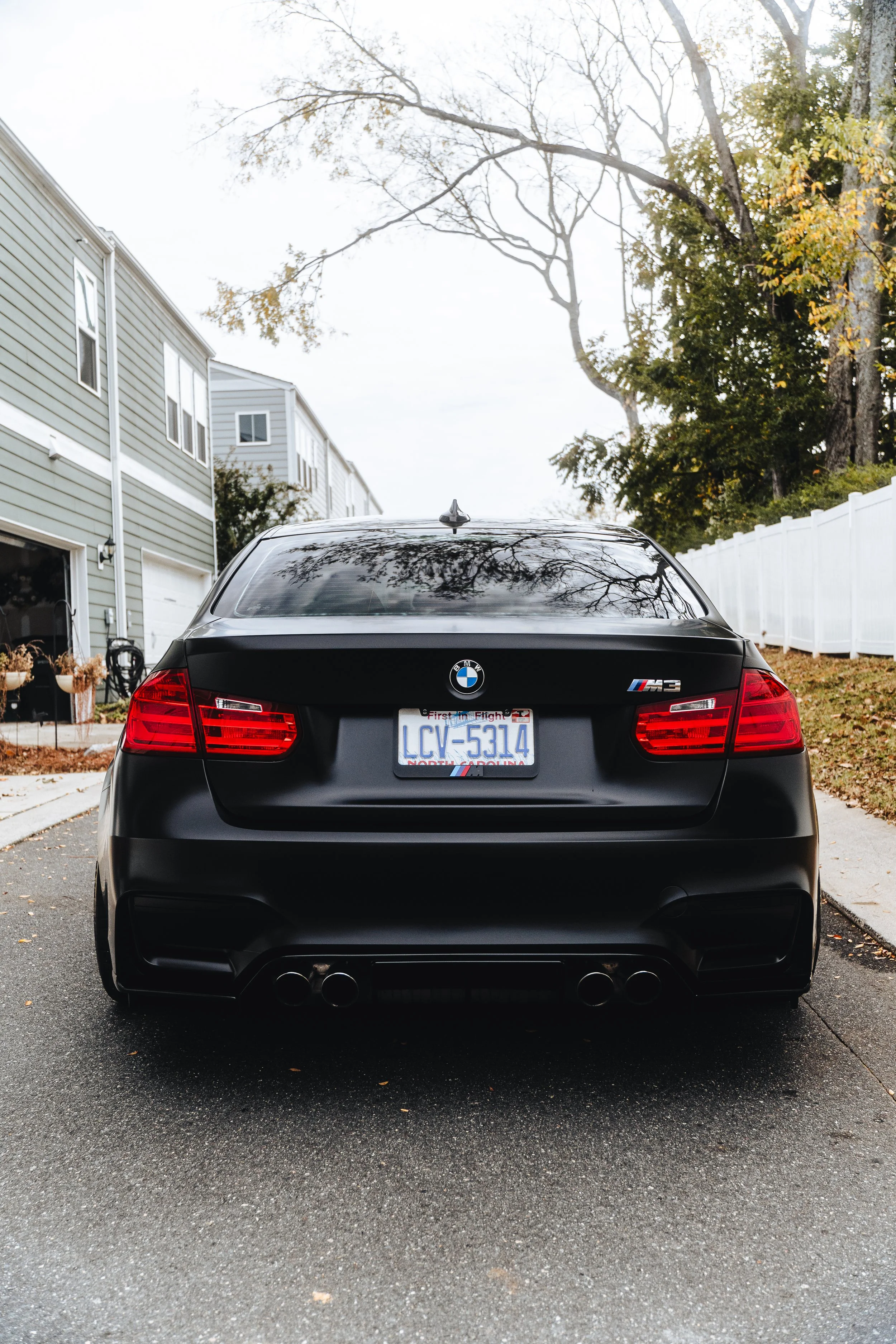 Black BMW M3 parked in a driveway with houses and trees in the background.