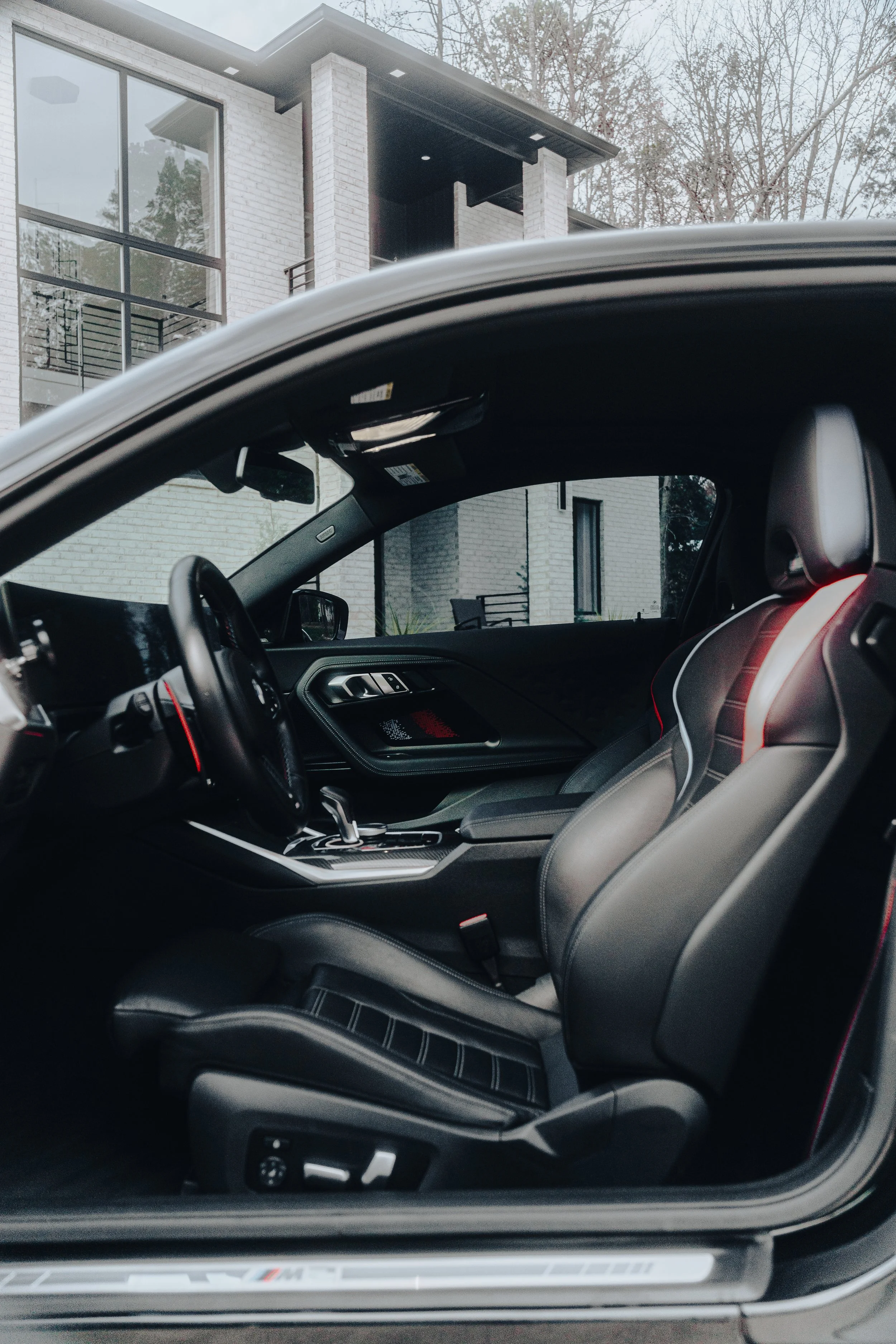 Inside view of a sports car with black leather seats, dashboard, and gear shift, parked in front of a modern white brick house.