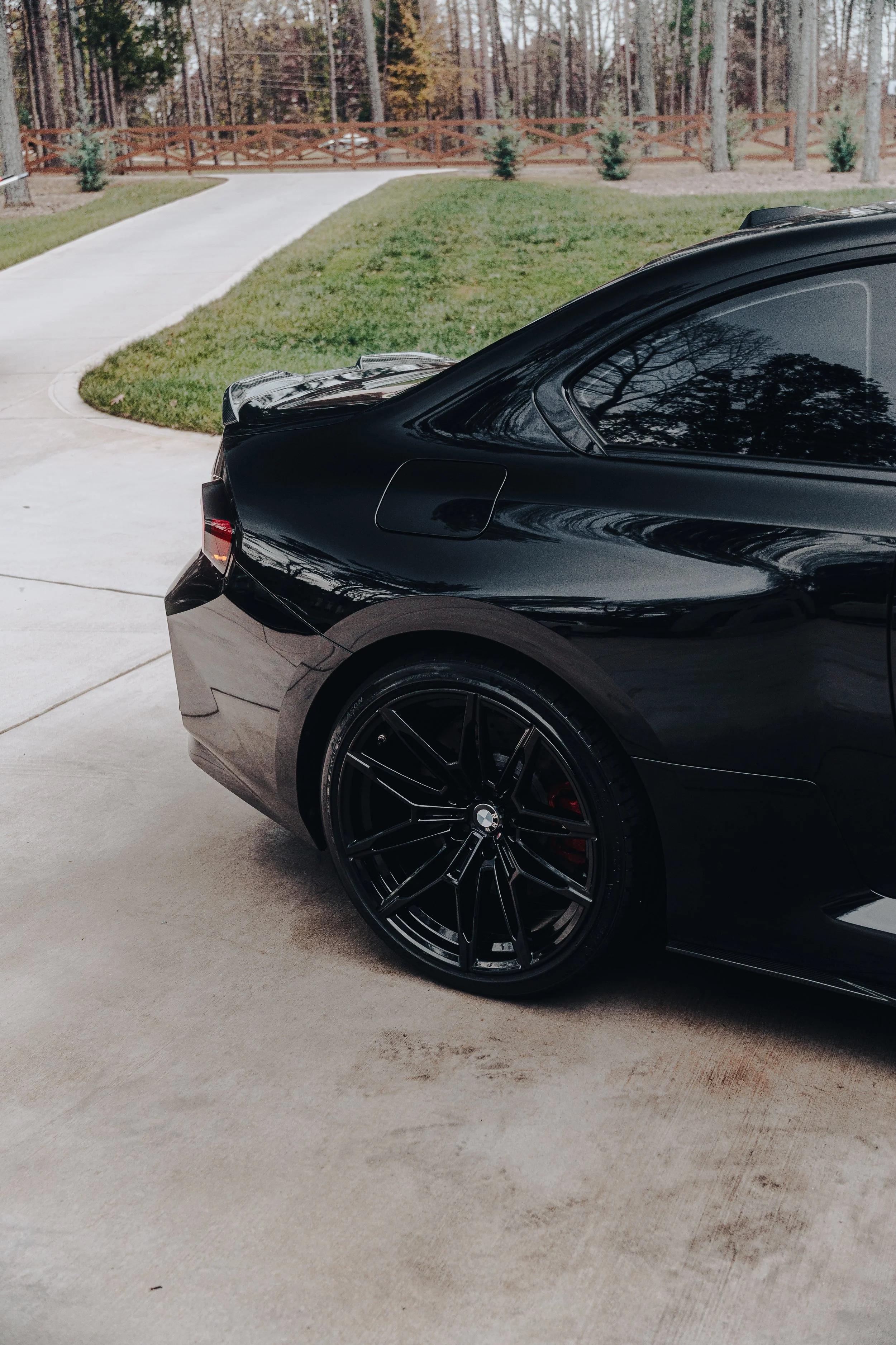 Close-up of the rear side of a black BMW sports car parked in a driveway, reflecting nearby trees and sky.