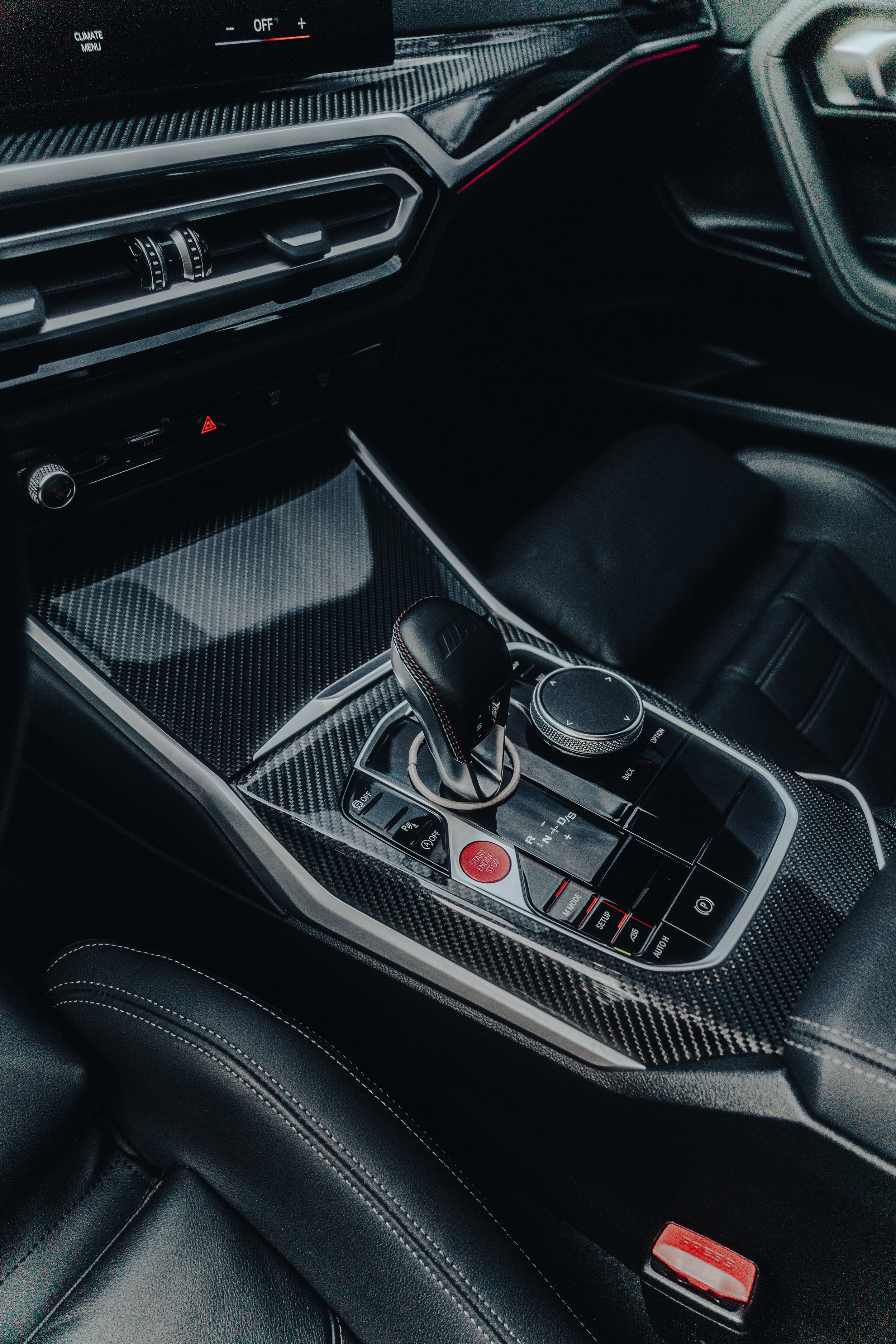 Close-up of a car's center console with a gear shifter, control buttons, and a rotary dial, featuring a carbon fiber finish.