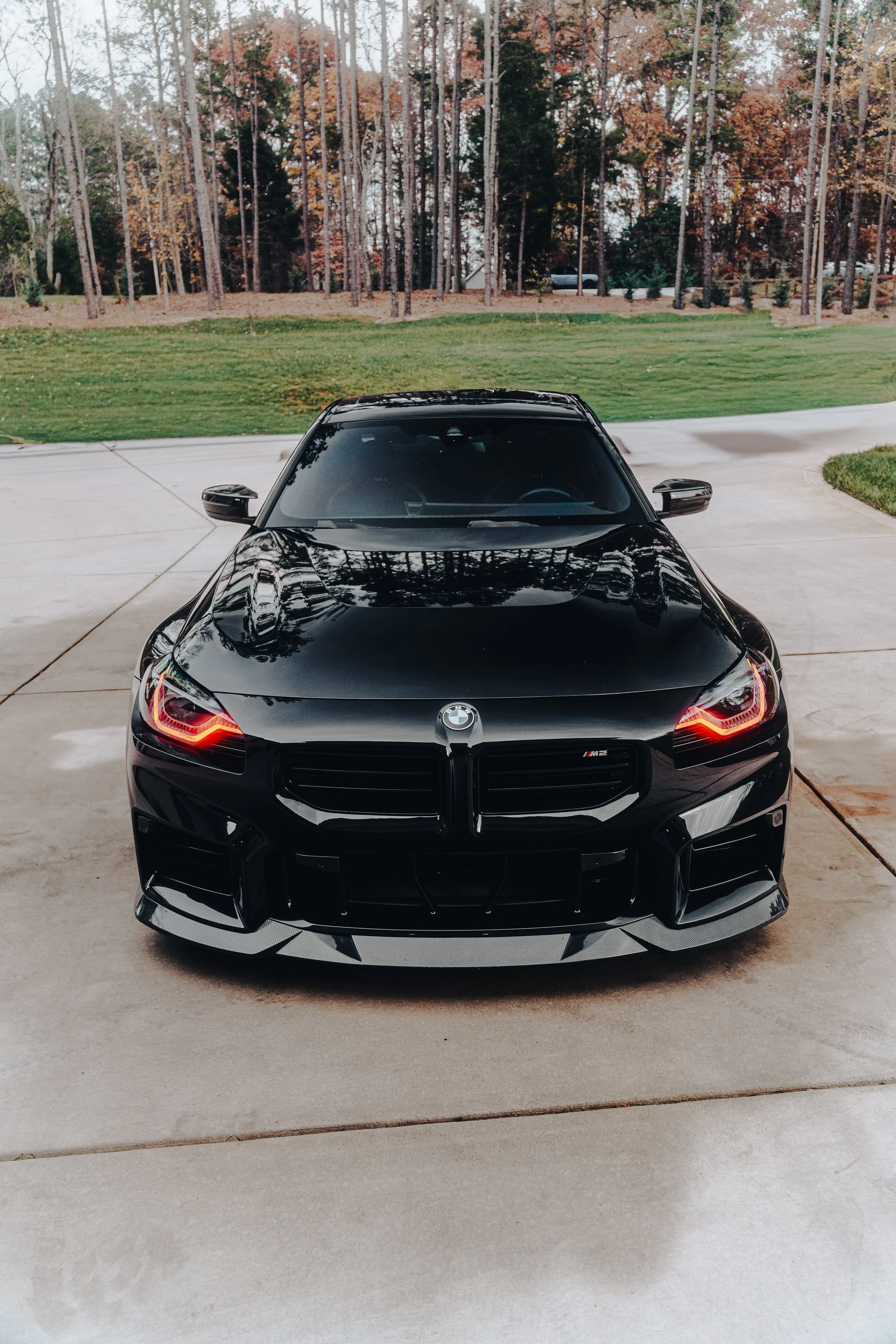 Front view of a black BMW M2 parked on a concrete driveway with trees and grass in the background.