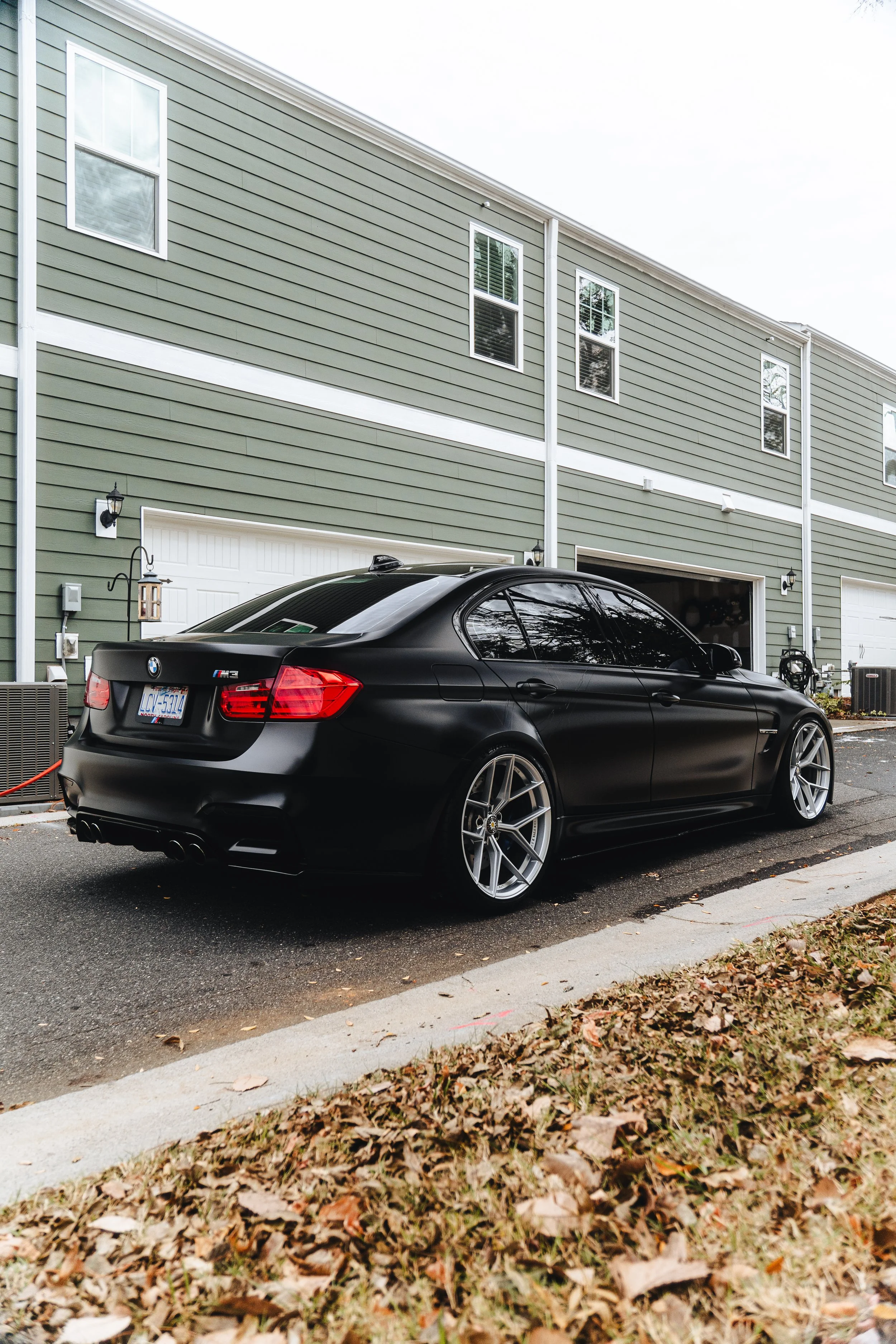 A black BMW M3 parked in a driveway next to a green residential building with white trim and three windows on the second floor.