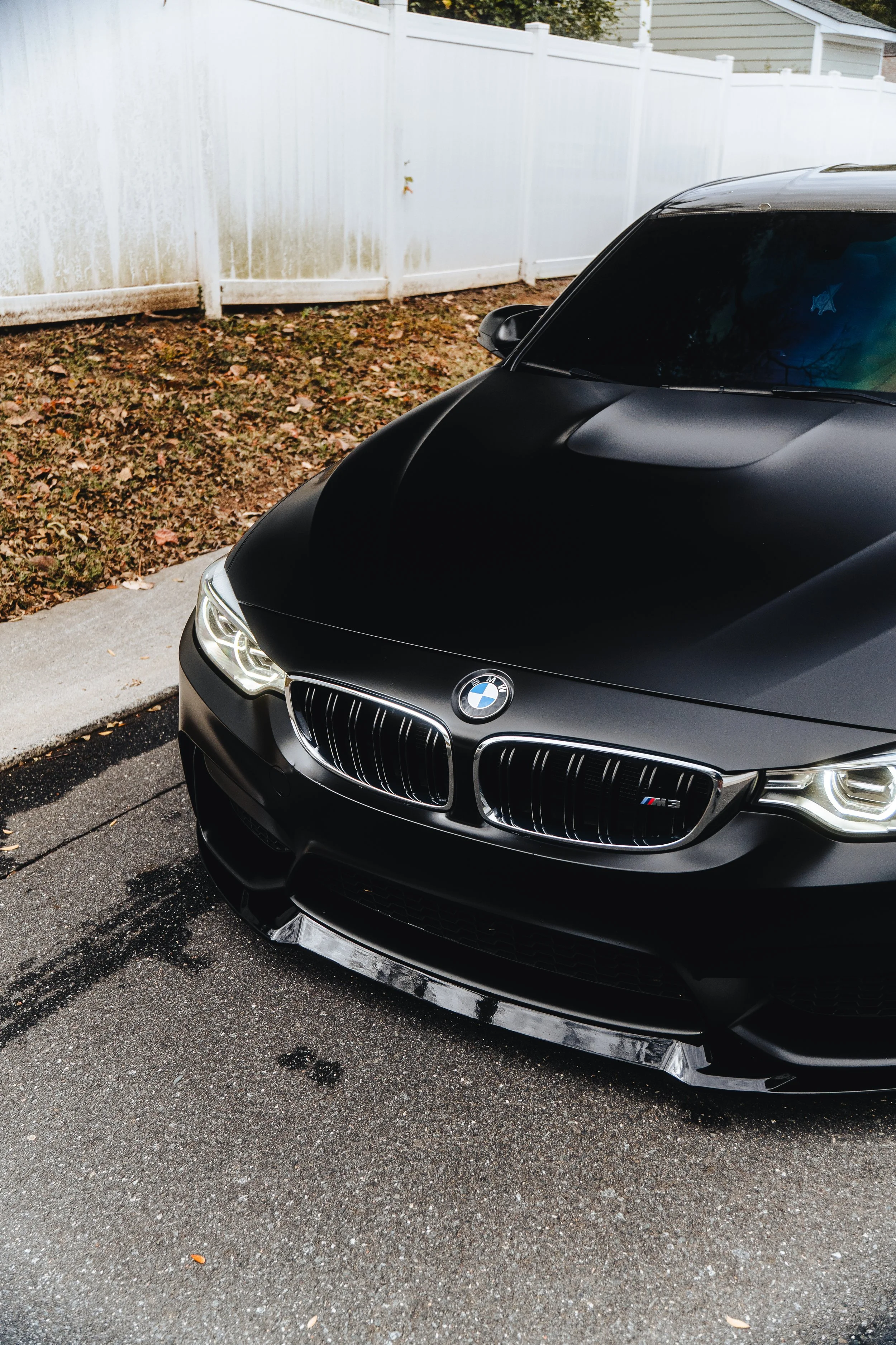 Black BMW car parked on a street near a sidewalk with dried leaves, with a white fence in the background.