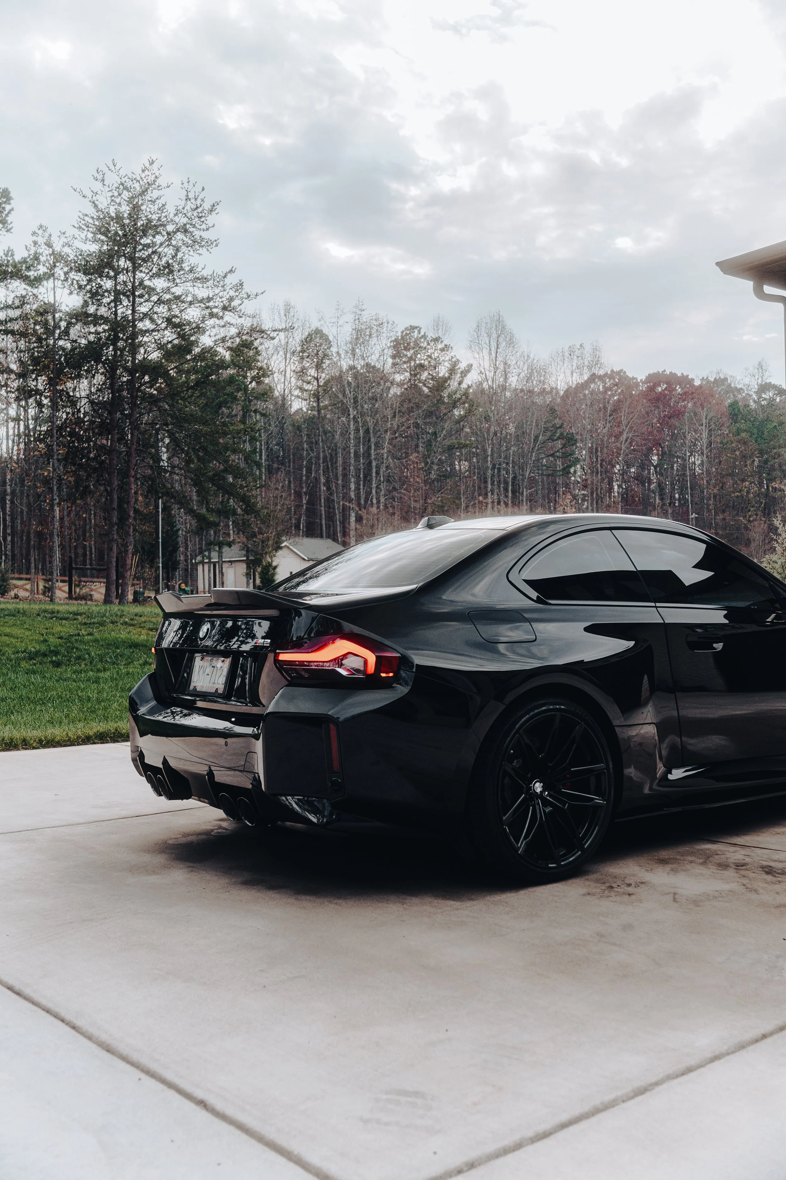 A sleek black sports car parked on a concrete driveway with a green lawn and trees in the background under a cloudy sky.
